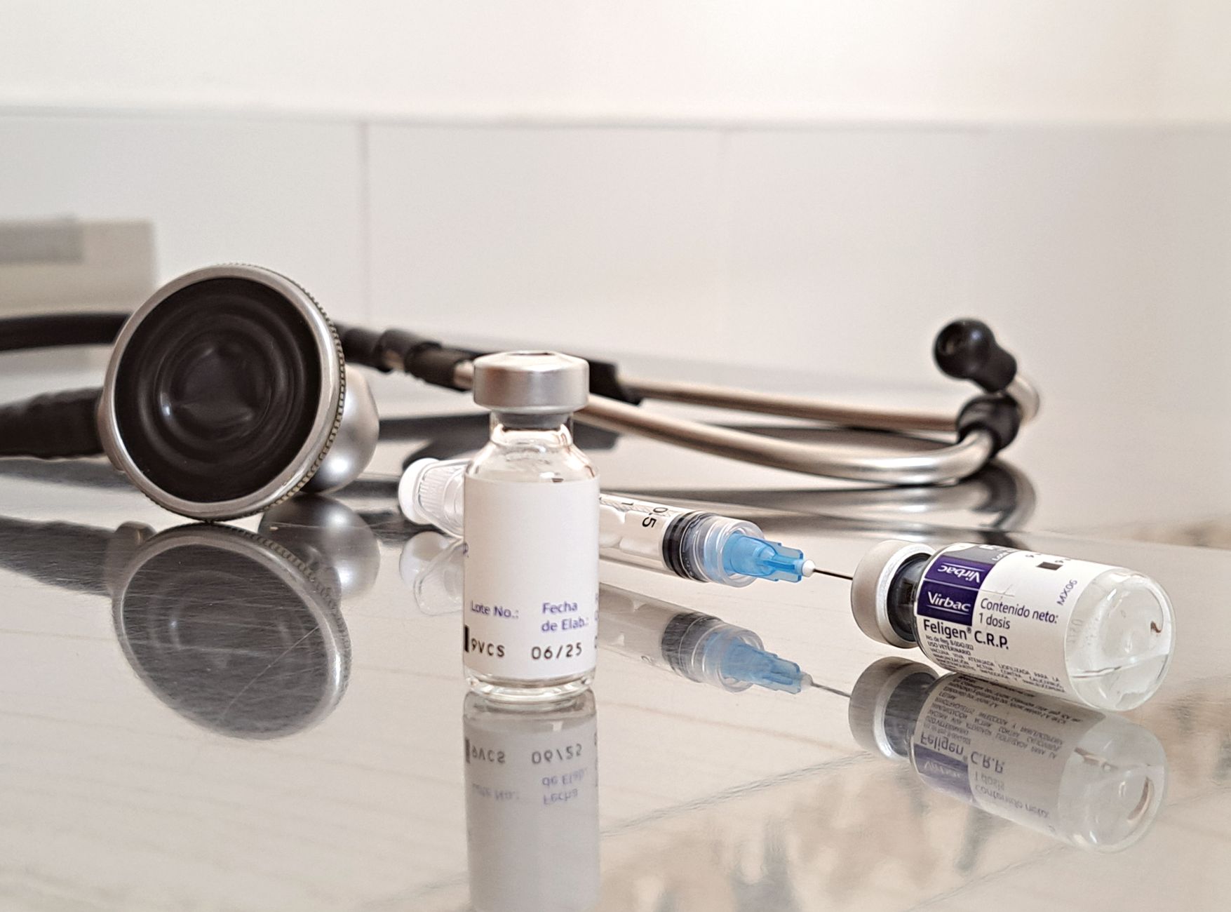 Close-up of vaccines, syringe, and stethoscope on a reflective surface in a clinical setting.