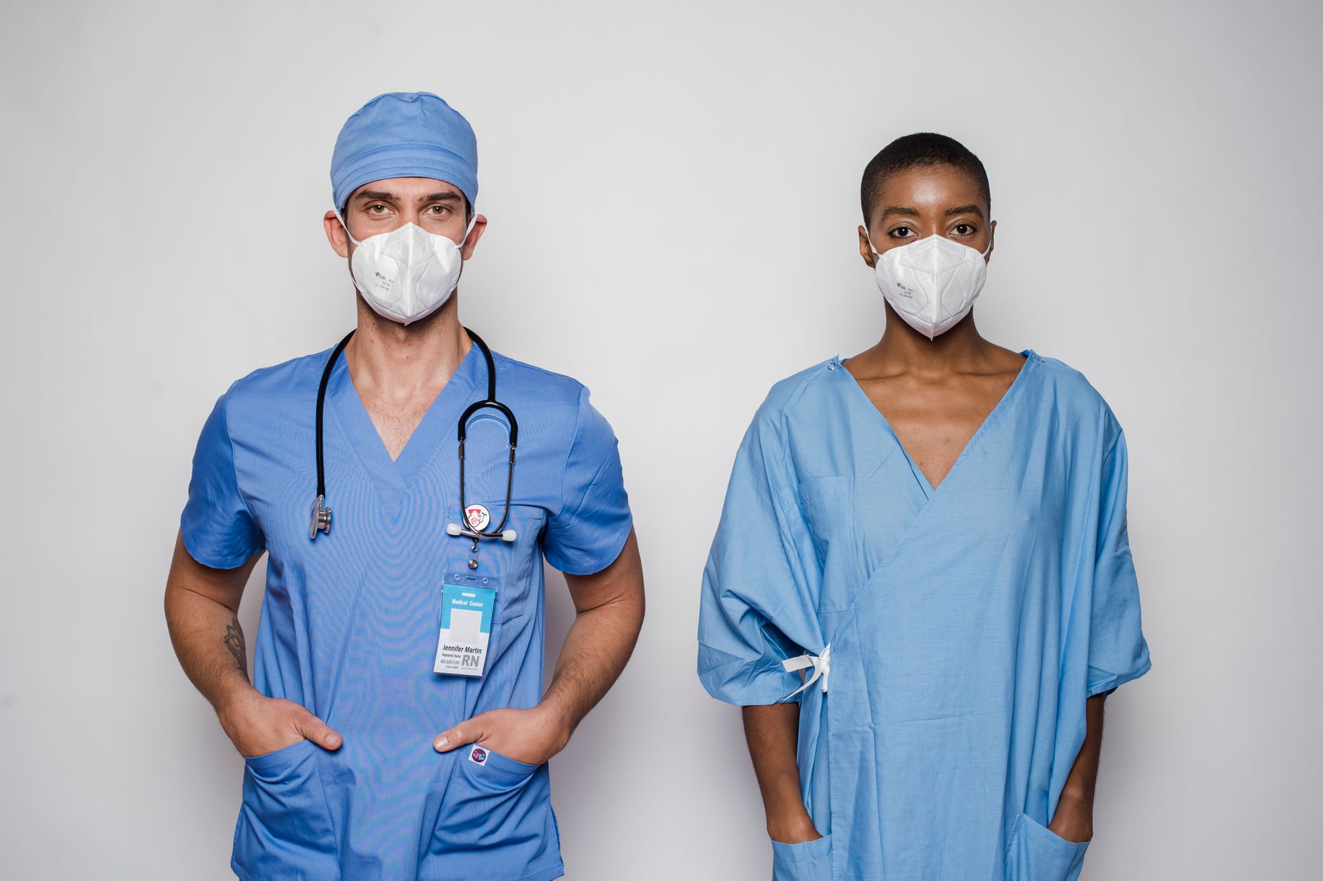 Multiethnic adult male doctor with stethoscope standing near female patient in blue uniform and protective masks in light studio on white background while looking at camera with hands in pockets