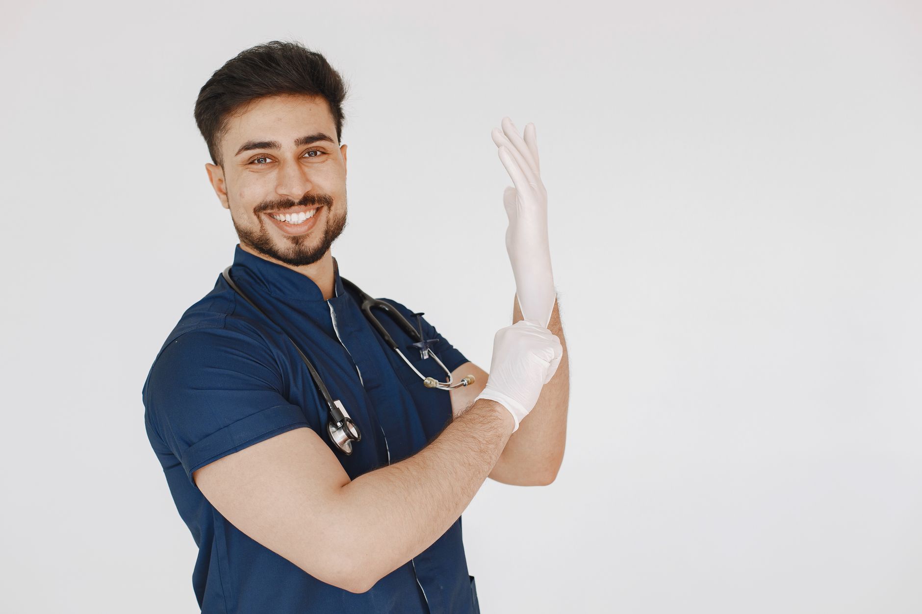 A male doctor in scrubs dons medical gloves, smiling confidently against a plain white background.