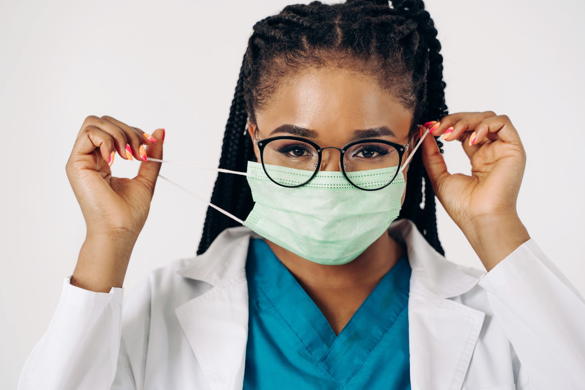 A female doctor adjusting her surgical mask, wearing scrubs and eyeglasses on a white background.