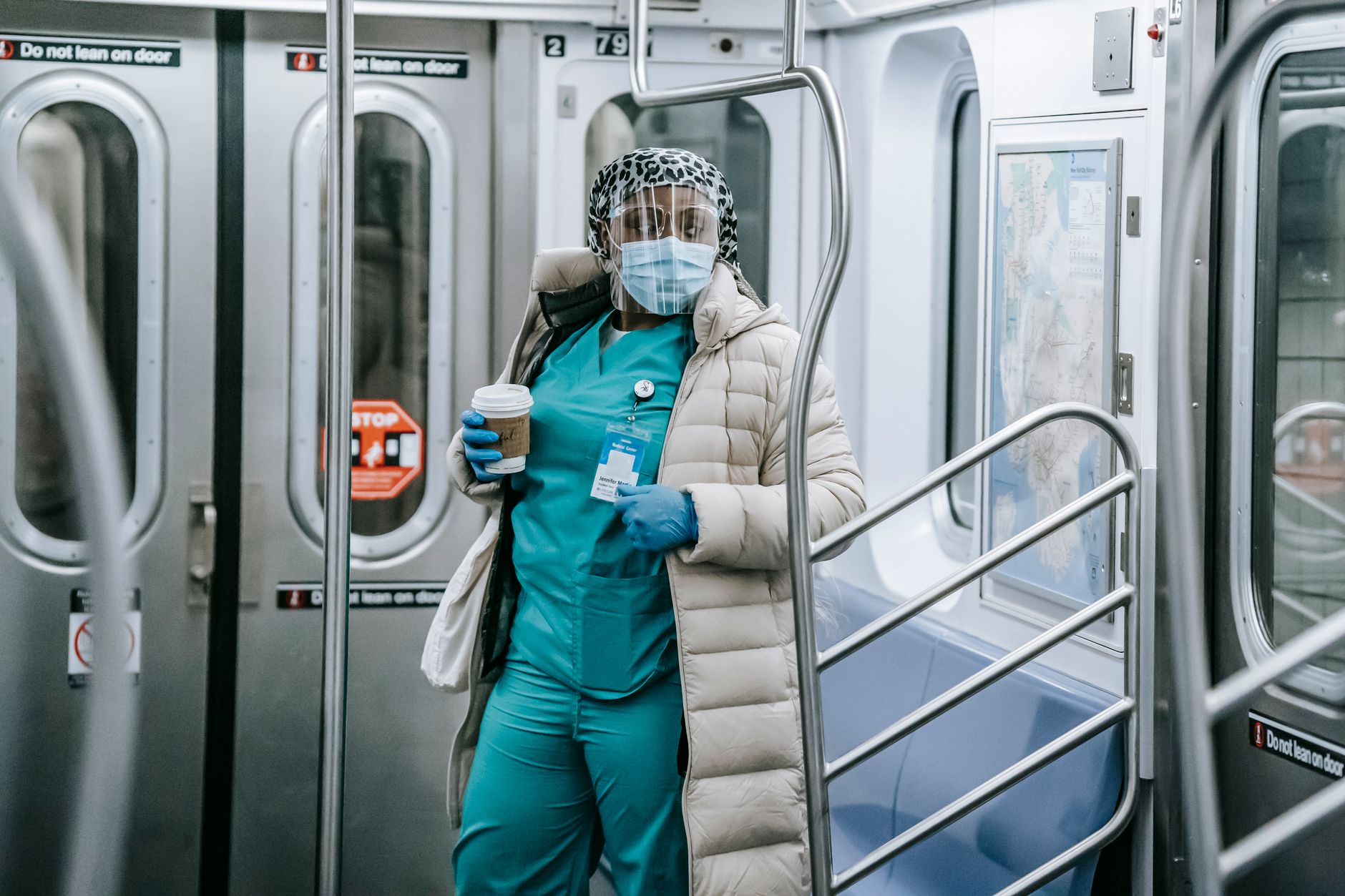 Calm African American female doctor in uniform under outerwear wearing protective mask and face shield standing with takeaway coffee in underground train