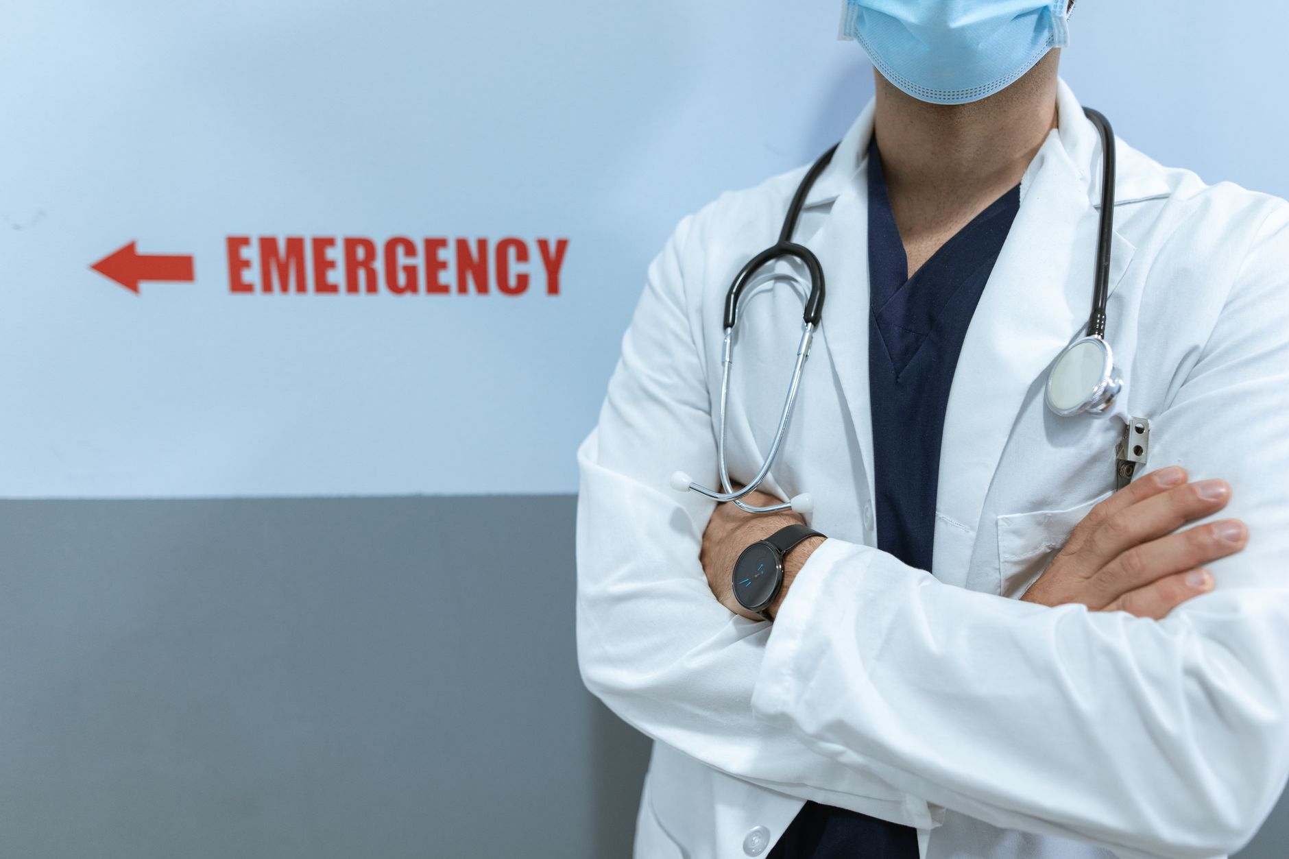 Healthcare worker in emergency room with stethoscope and facemask, cross-armed posture.