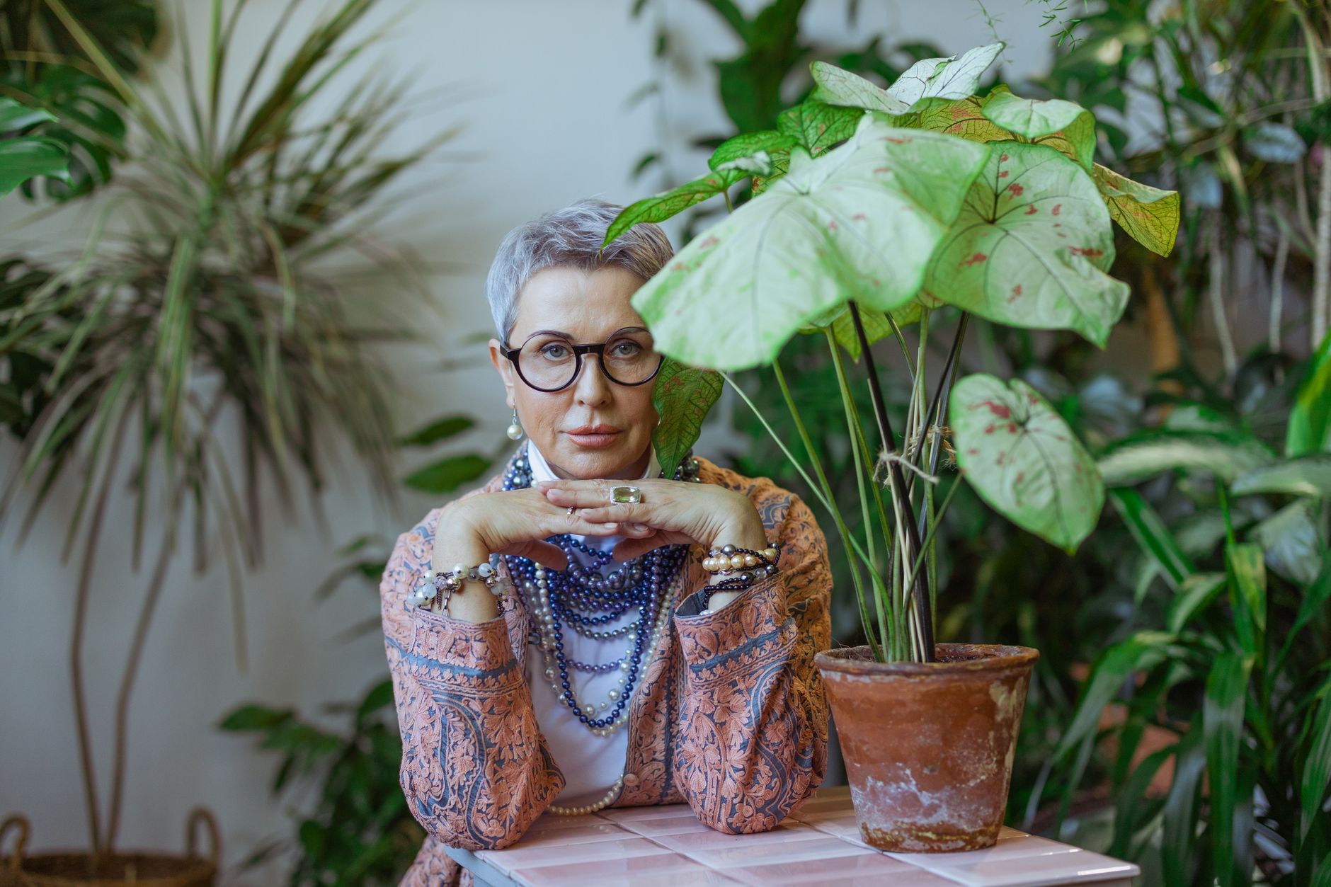 Elegant woman with glasses and gray hair, posing with hand resting on a table surrounded by lush indoor plants.