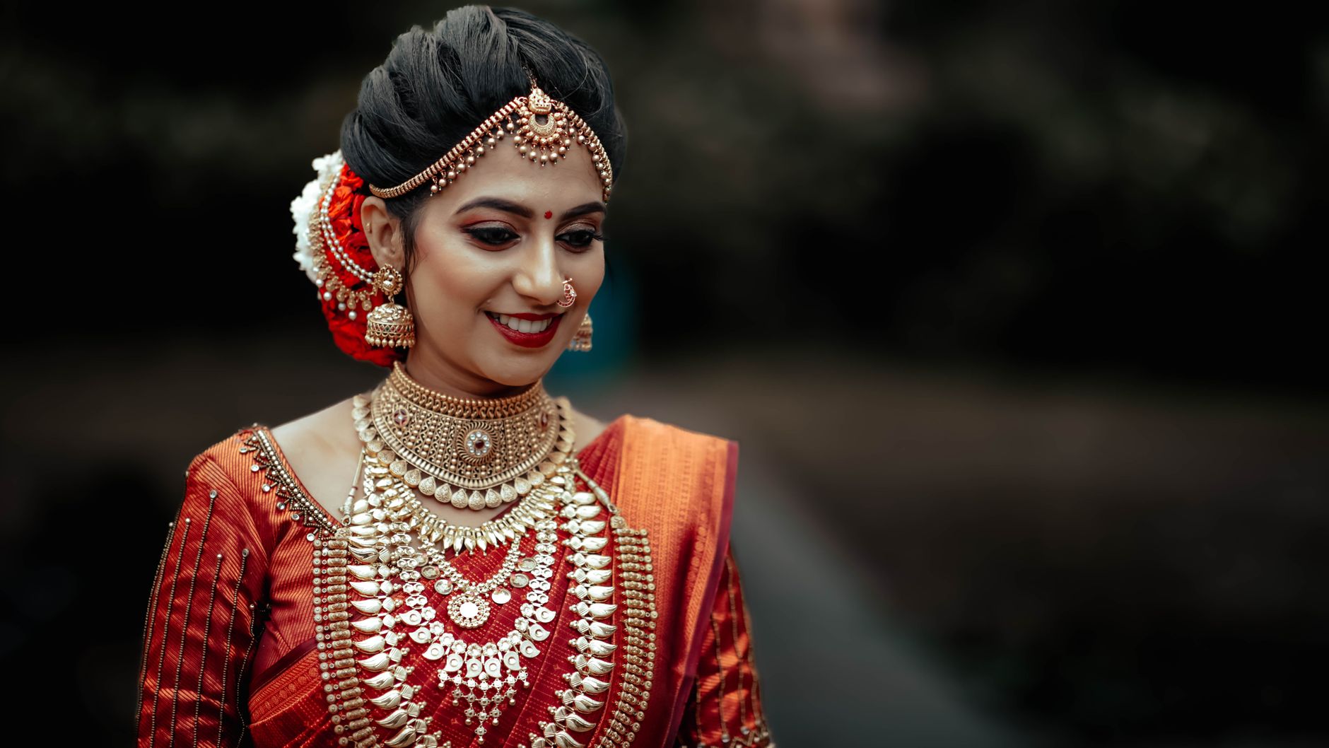 South Asian woman in rich traditional bridal attire with intricate jewelry and sari, smiling gently.