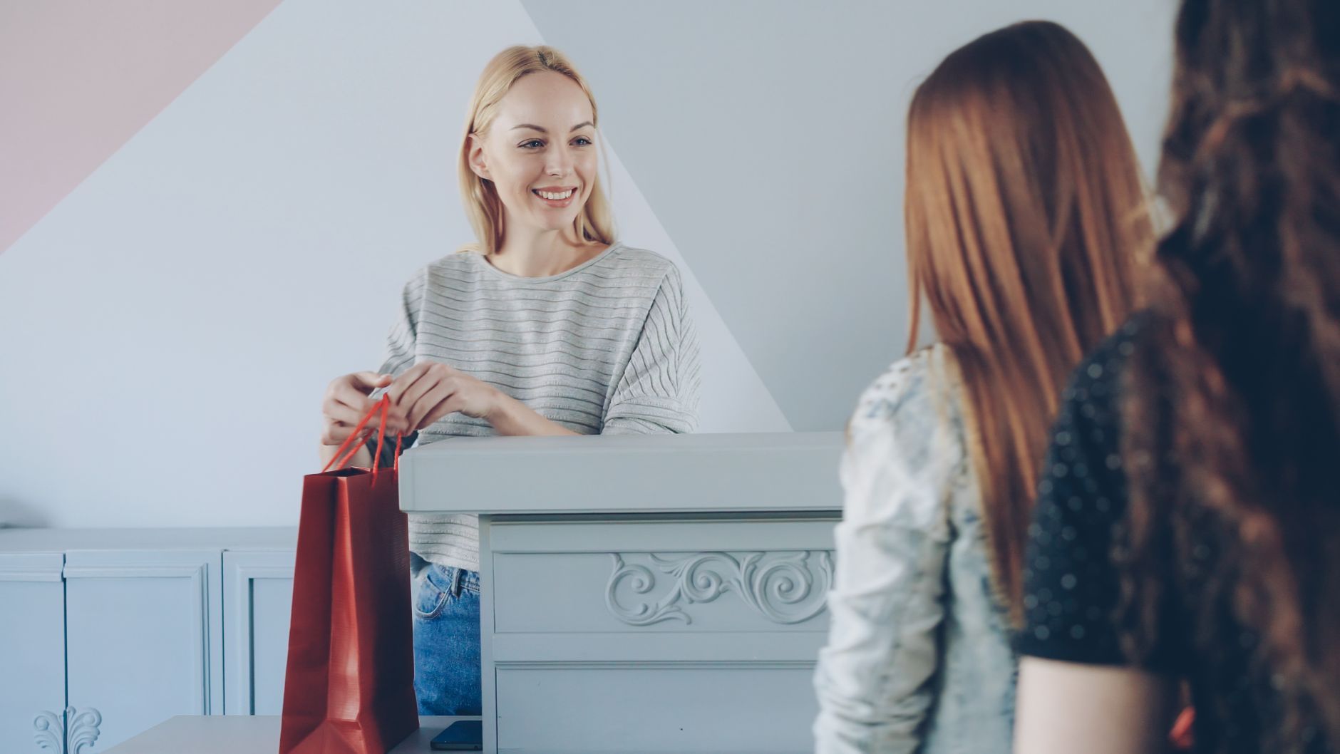 A cheerful cashier hands a red shopping bag to two customers in a stylish store.