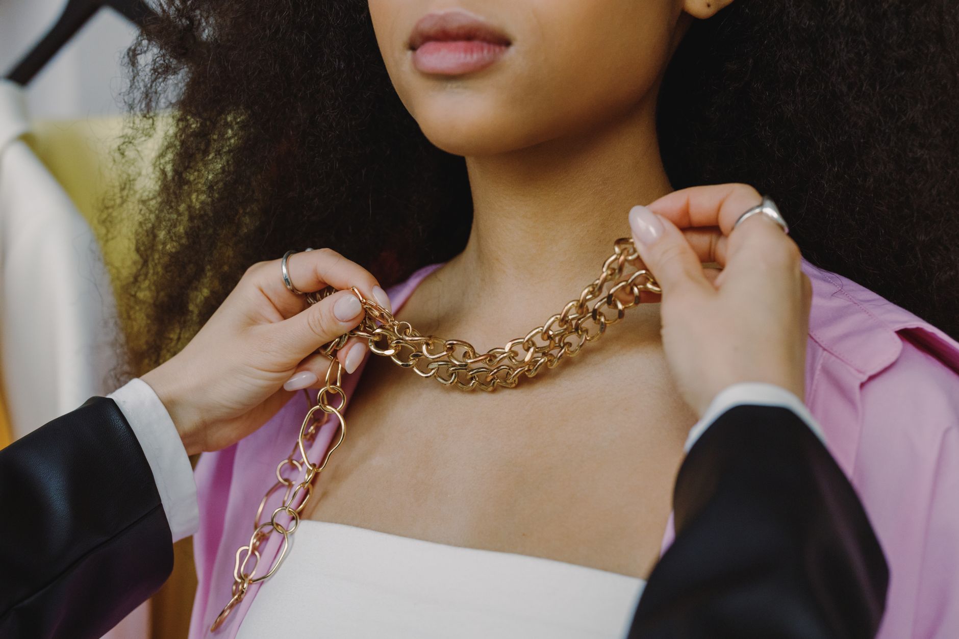 Close-up of a stylist adjusting a gold chain necklace on a model wearing a pink blazer indoors.