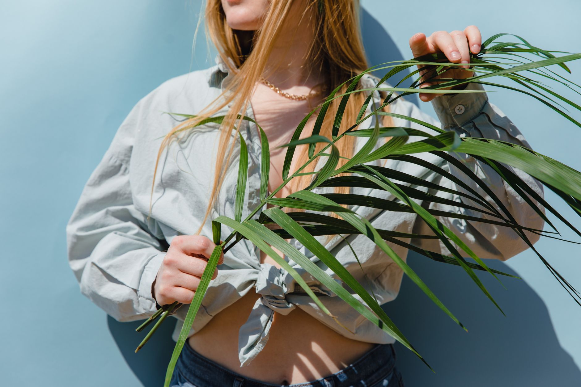 Fashionable woman in casual shirt holding a palm leaf with blue background, showcasing modern style.