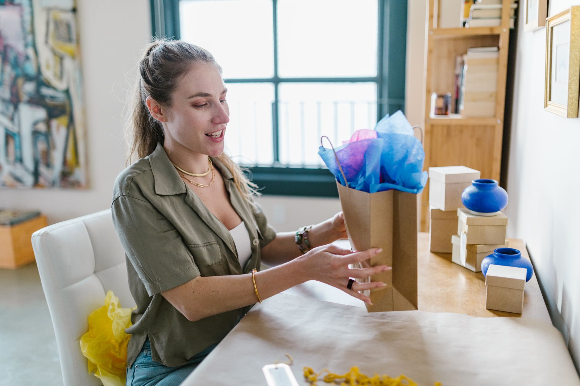Woman sitting at a table wrapping a gift with colorful tissue paper and craft supplies.