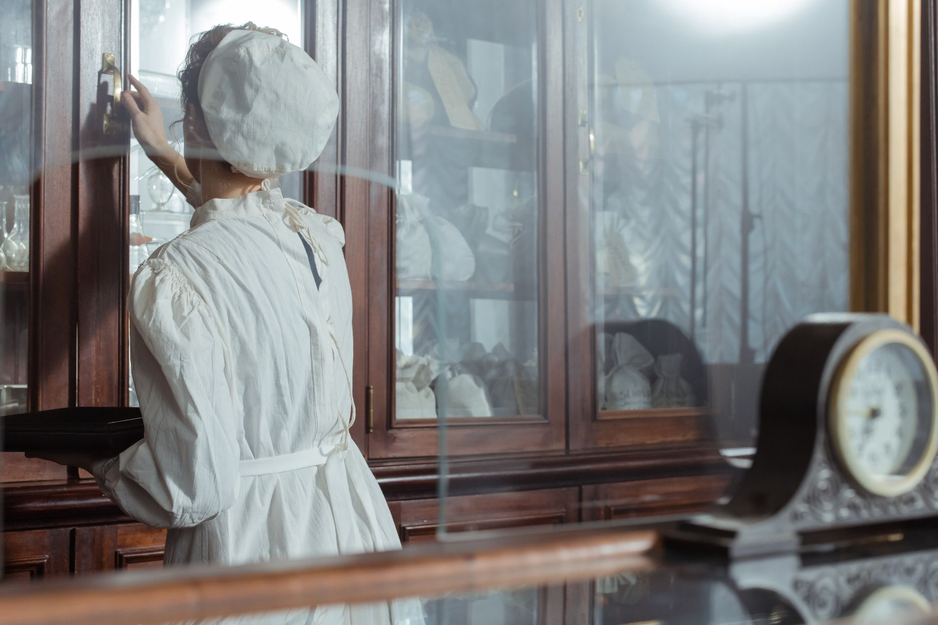 A woman in vintage attire retrieves items from a pharmacy cabinet.
