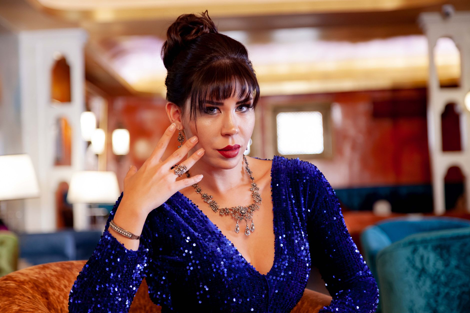 Sophisticated woman posing indoors in a luxurious blue sequin dress and ornate jewelry, exuding elegance.