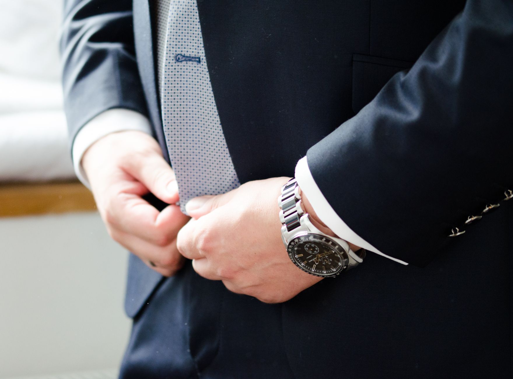 Close-up of groom adjusting tie, wearing a formal suit with watch, indoors.