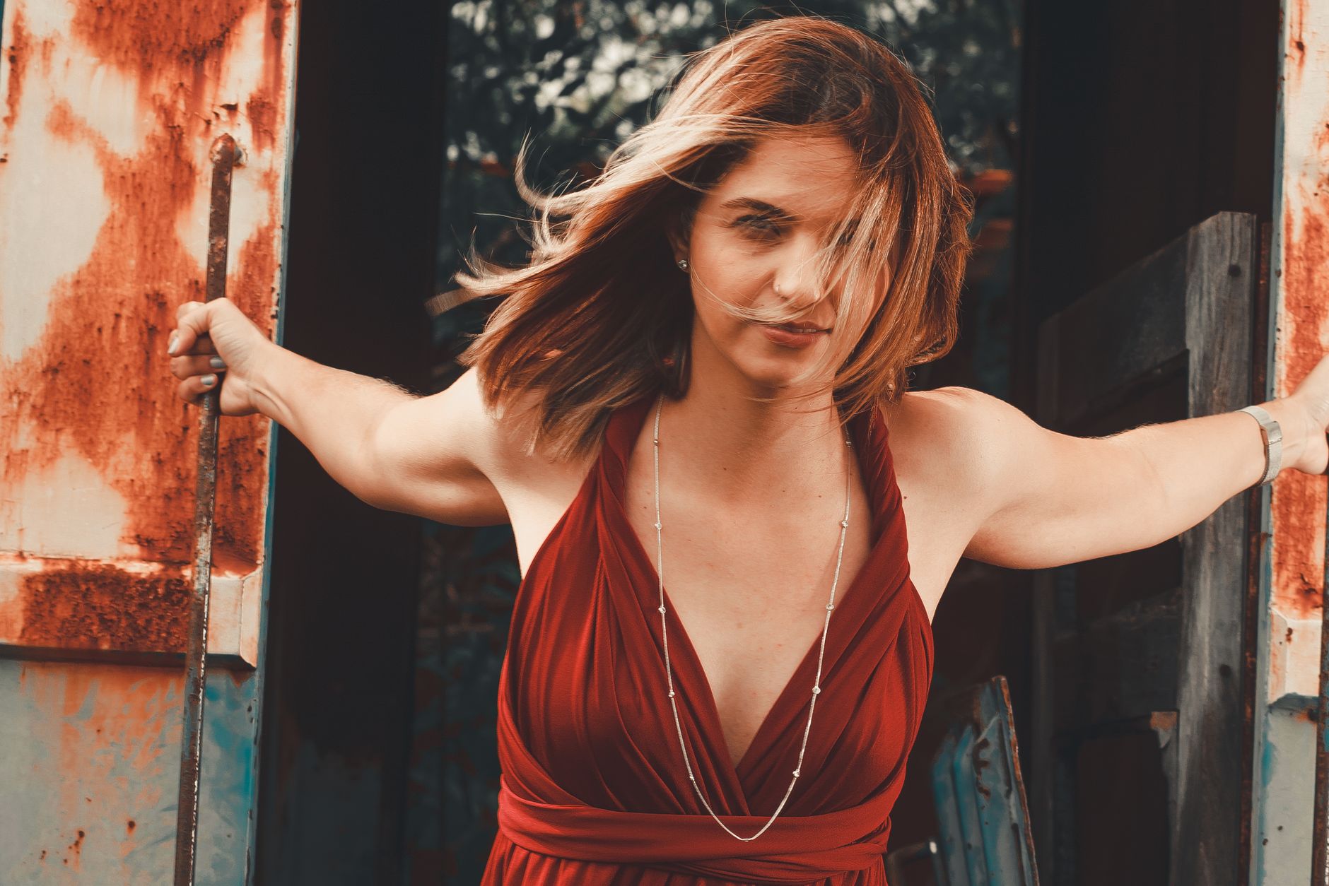 Elegant woman in red gown poses confidently at a rusty urban backdrop, capturing summer vibes.