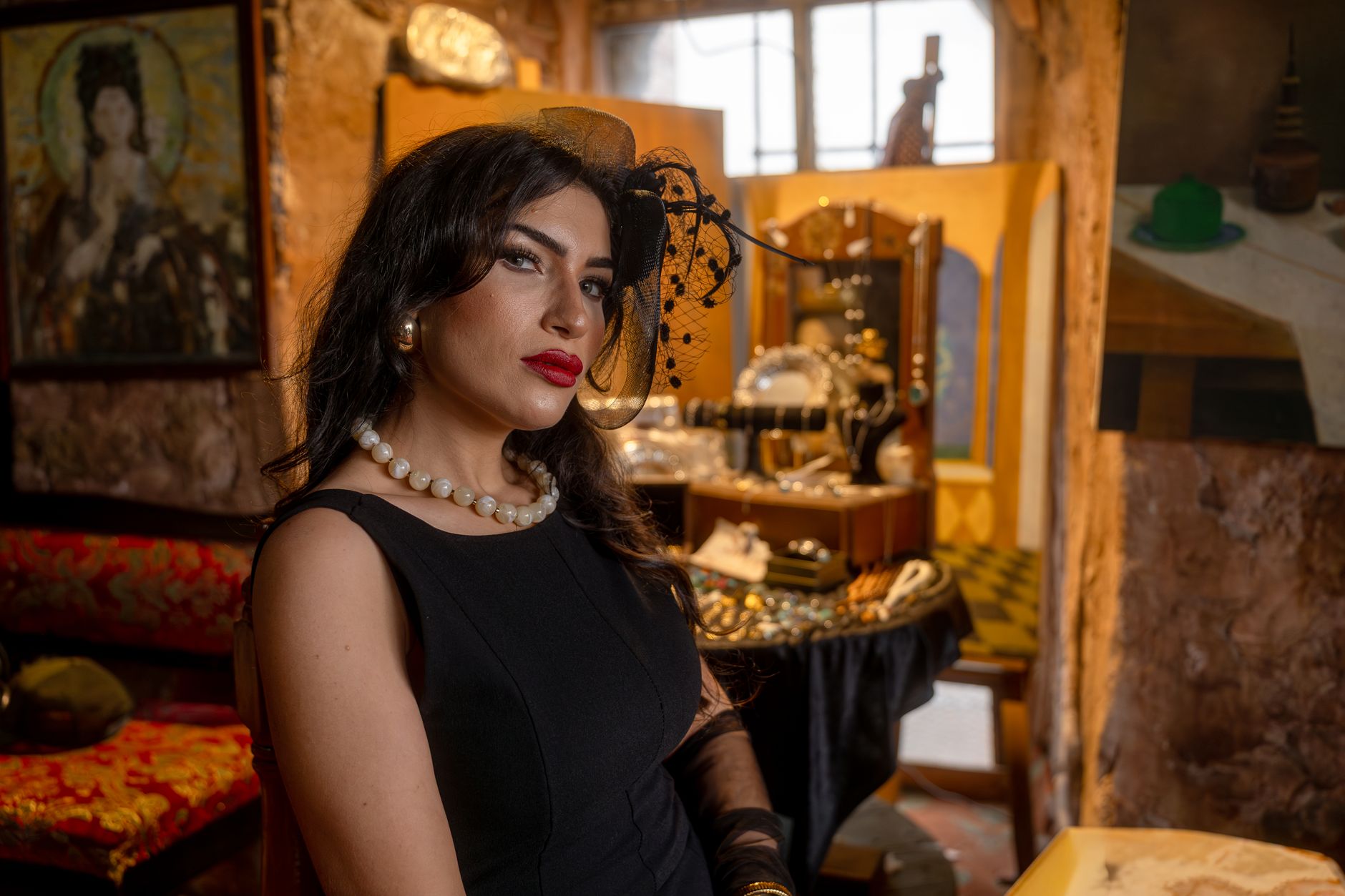 Sophisticated woman in black dress with pearl necklace in a vintage setting, surrounded by jewelry on display.
