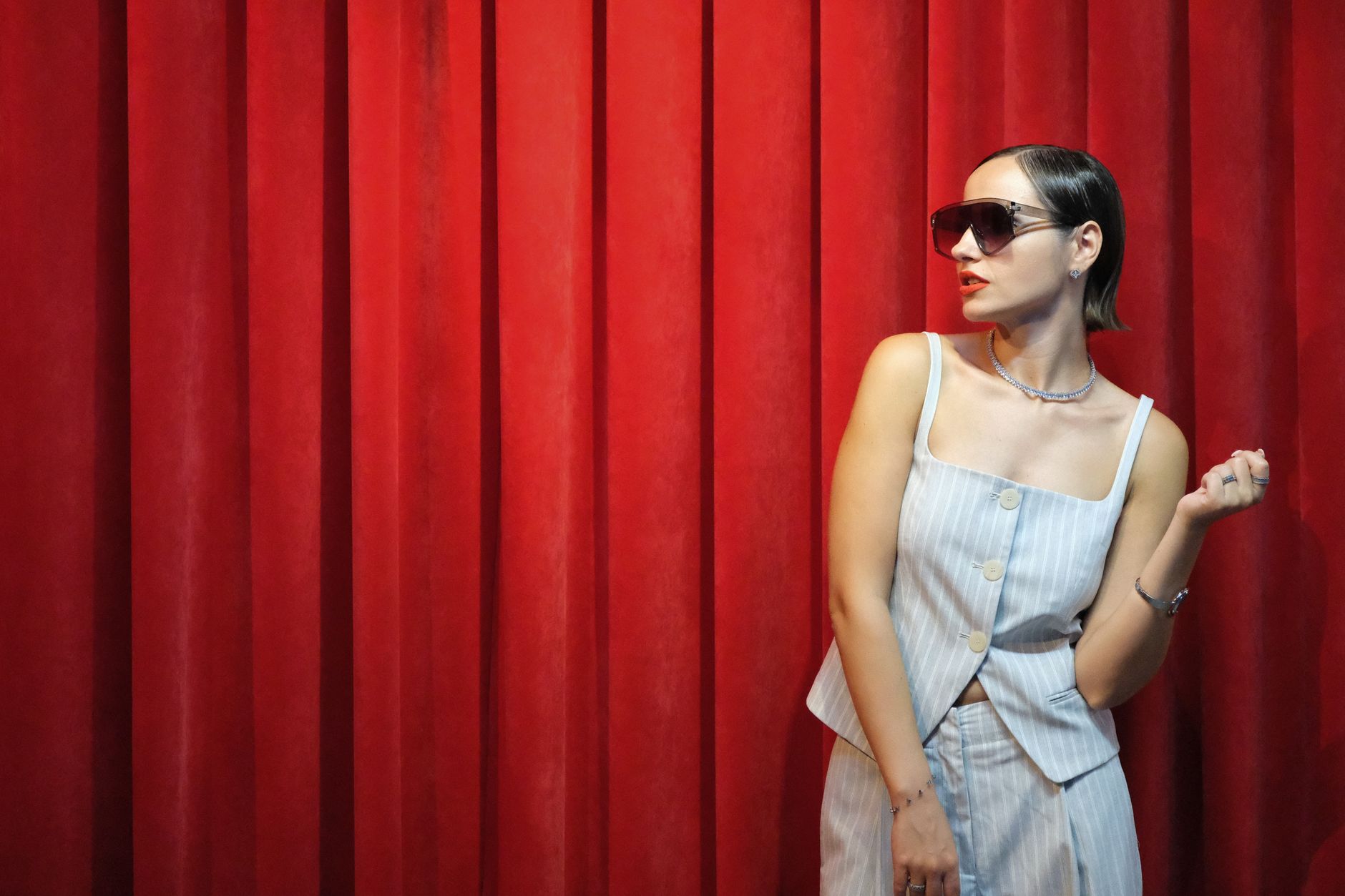 Chic woman in sunglasses poses against vibrant red curtains, exuding elegance.