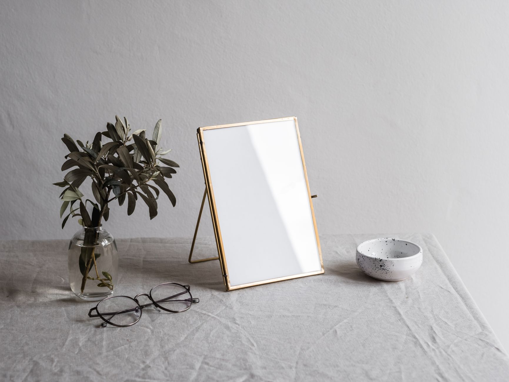 Elegant minimalist still life featuring a blank frame, eyeglasses, a bowl, and a leafy vase on a linen-covered surface.