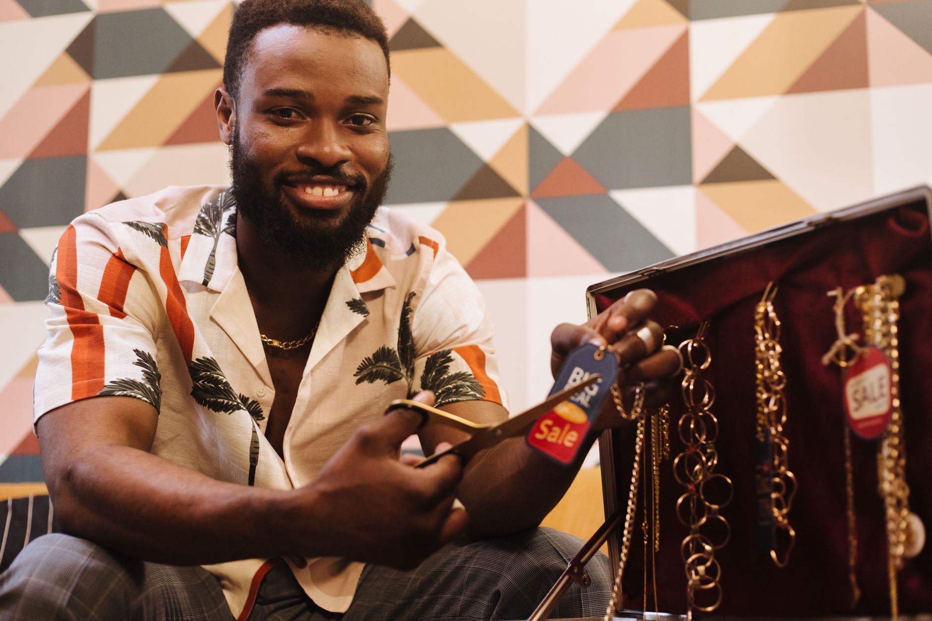 African American man smiling, holding a sale tag and showcasing jewelry in a colorful indoor setting.