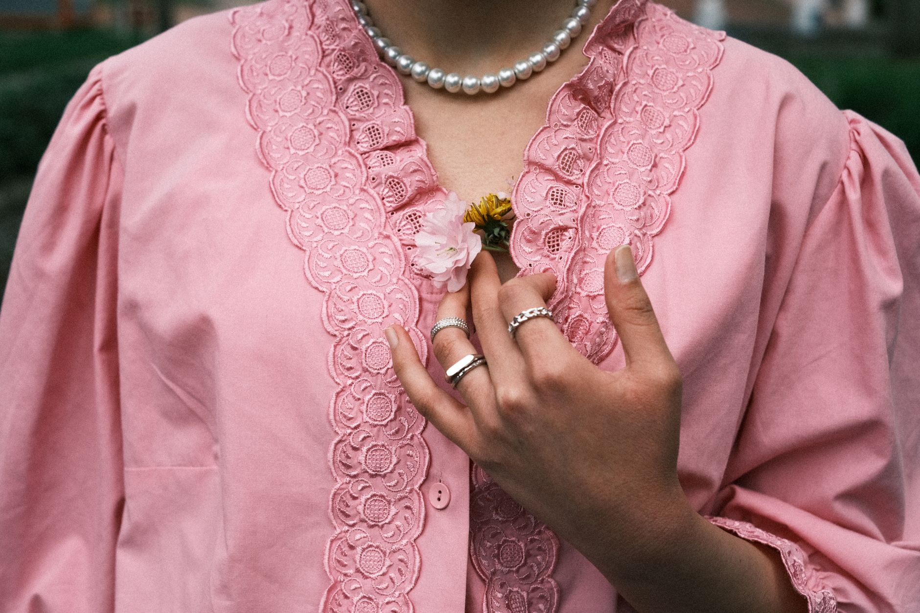 Close-up of a hand holding flowers on a pink lace blouse with pearl necklace.