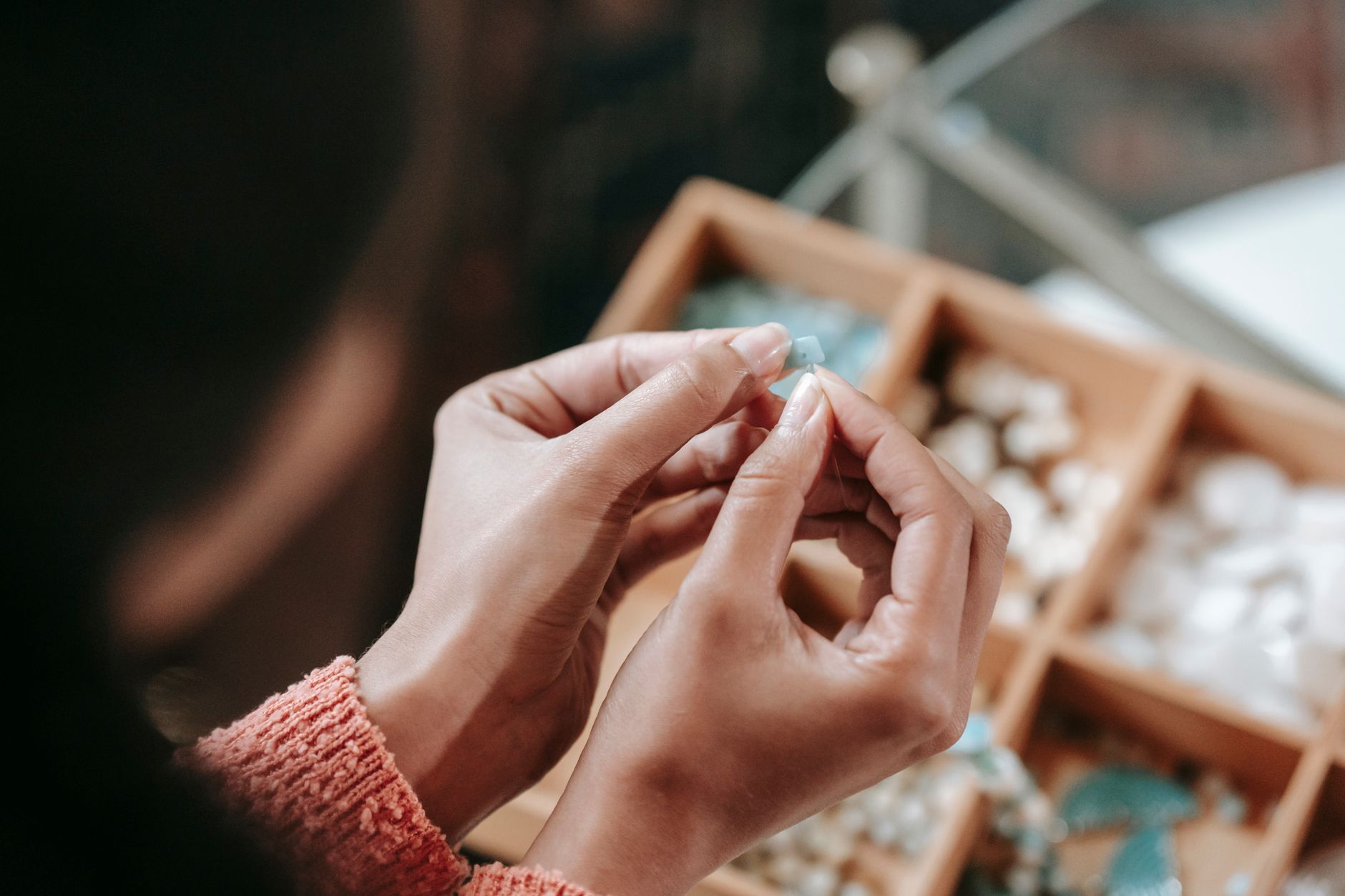 From above of crop faceless female stringing colorful bead on elastic rope while sitting at table with container of various supply