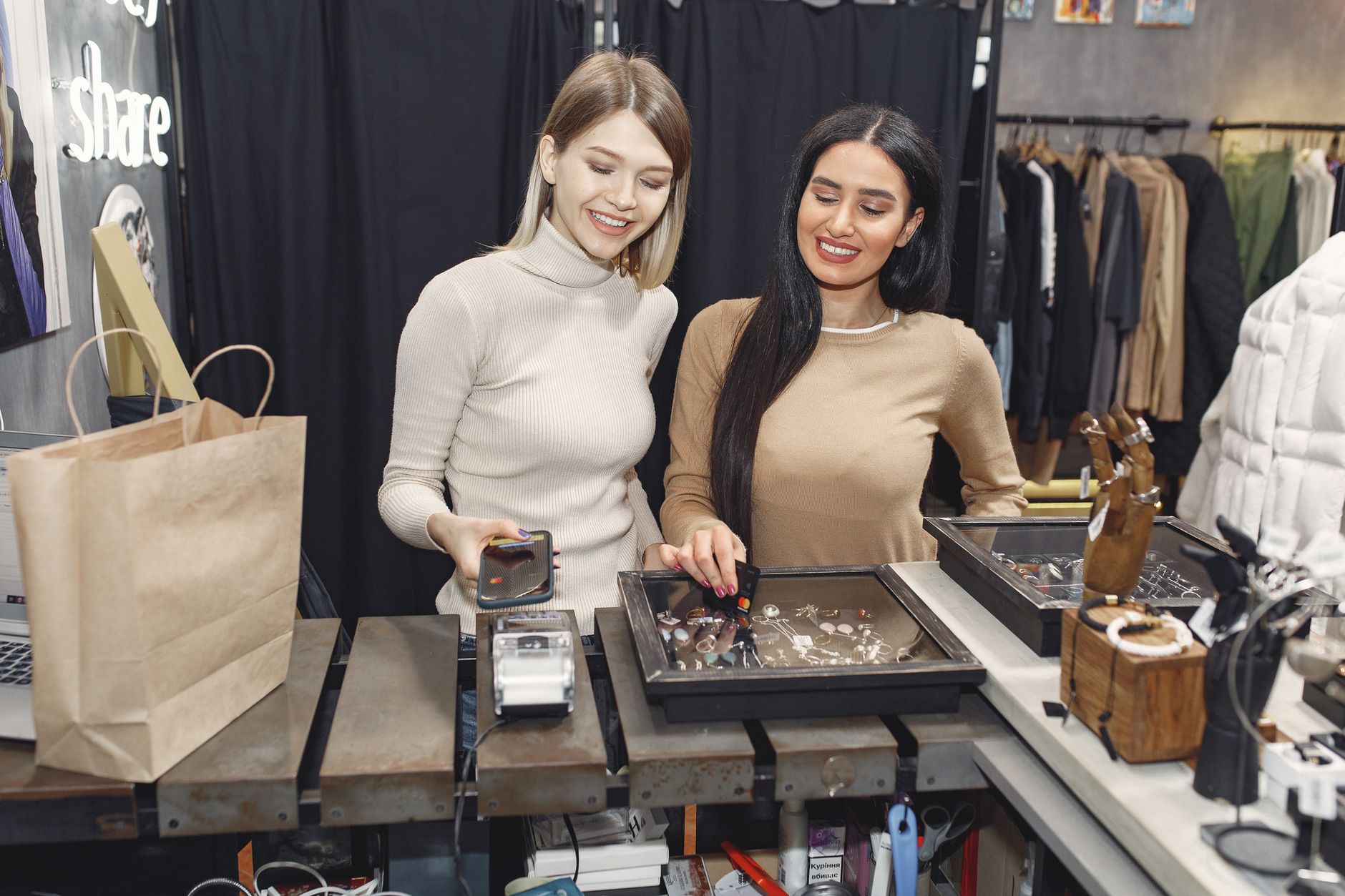 Cheerful casually dressed women paying with smartphone and credit card for purchases while standing near contactless terminal at counter desk