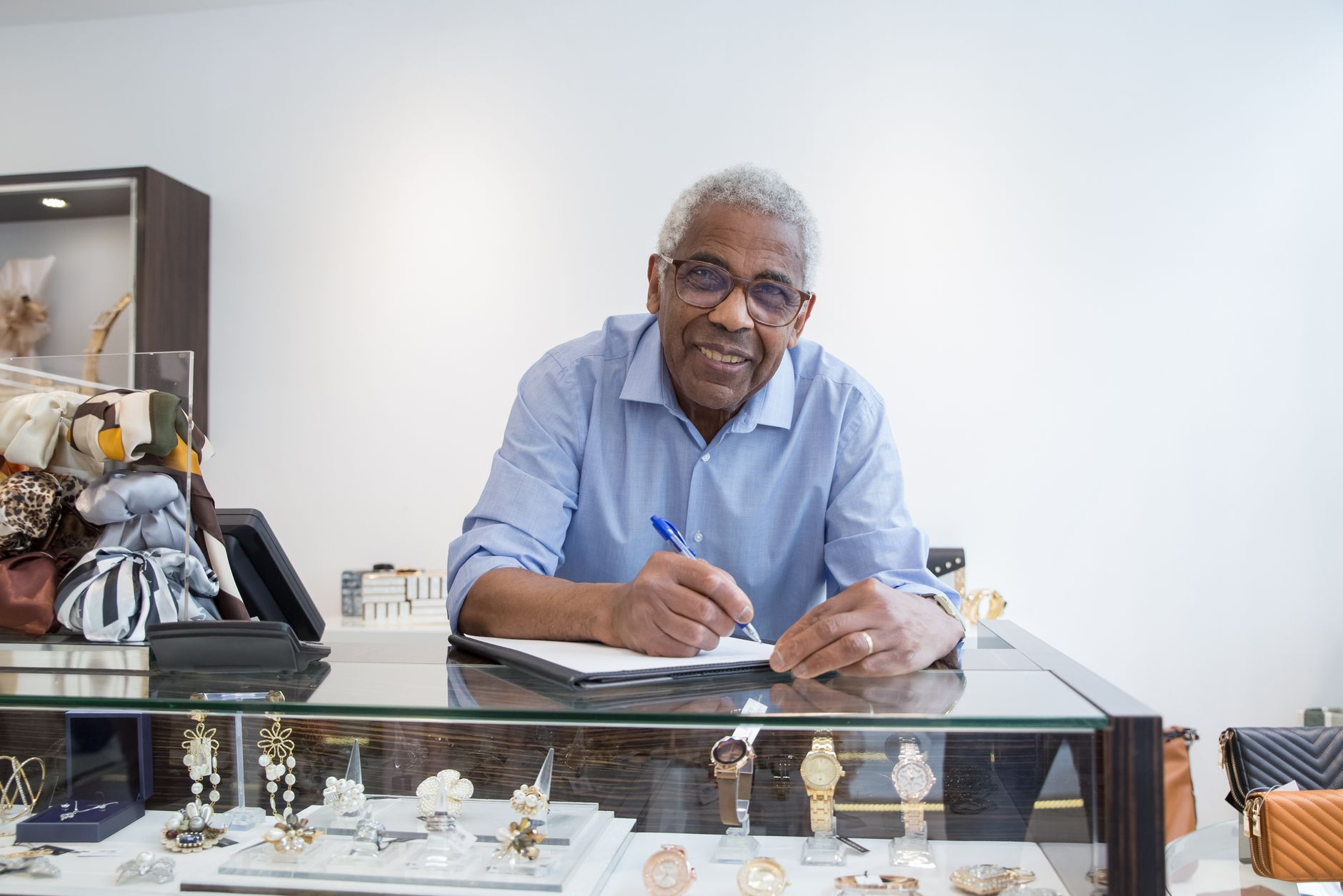 Smiling elderly shopkeeper in Portugal writing at jewelry display counter with eyeglasses.