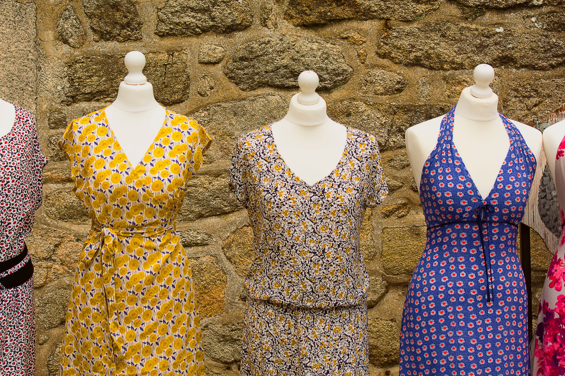 Vibrant floral dresses displayed on mannequins against a stone wall.