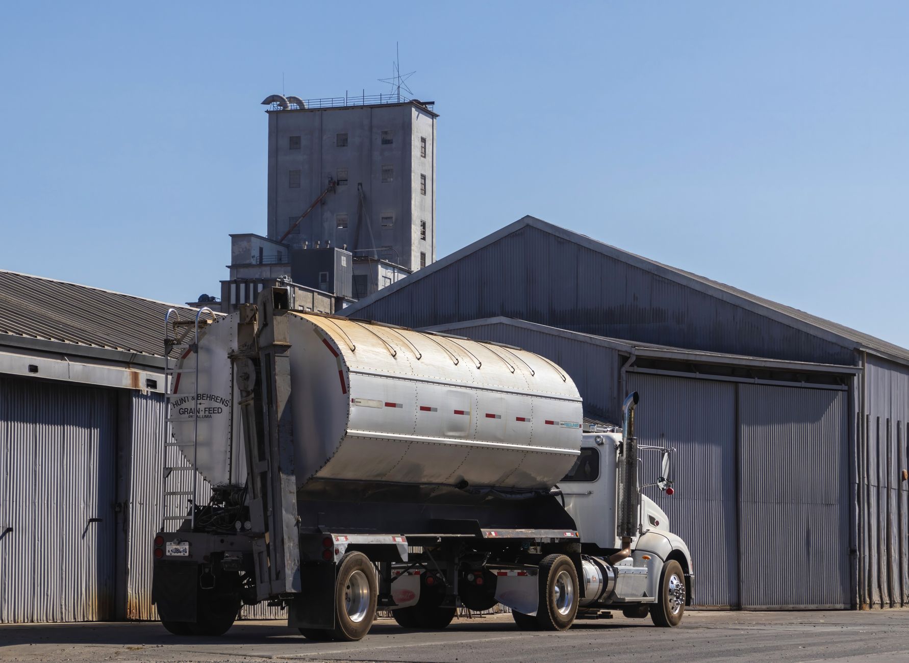 A large semi-trailer truck parked at an industrial grain warehouse under clear skies.
