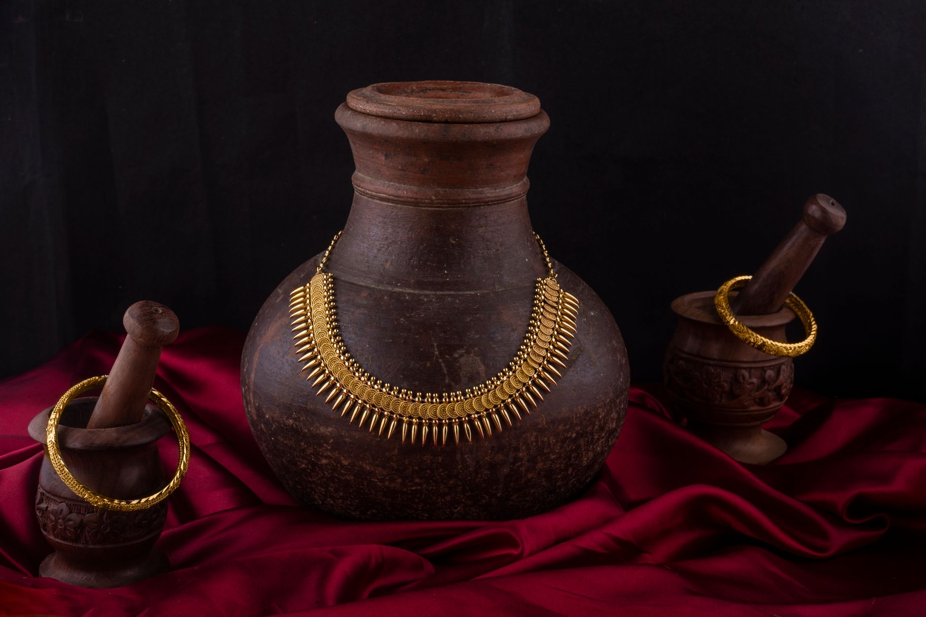 Elegant still life of traditional Indian necklace and bangles on vintage pottery with red fabric backdrop.