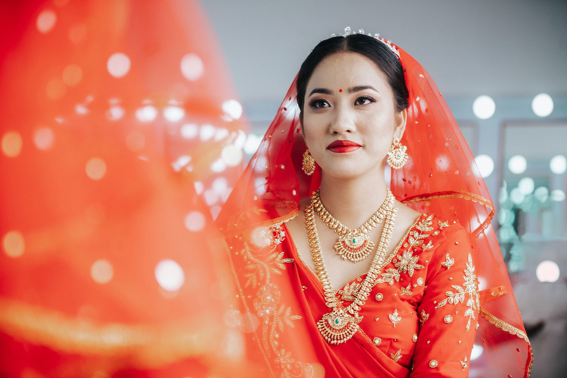 A young South Asian bride in vibrant red traditional attire, adorned with jewelry, smiling elegantly.