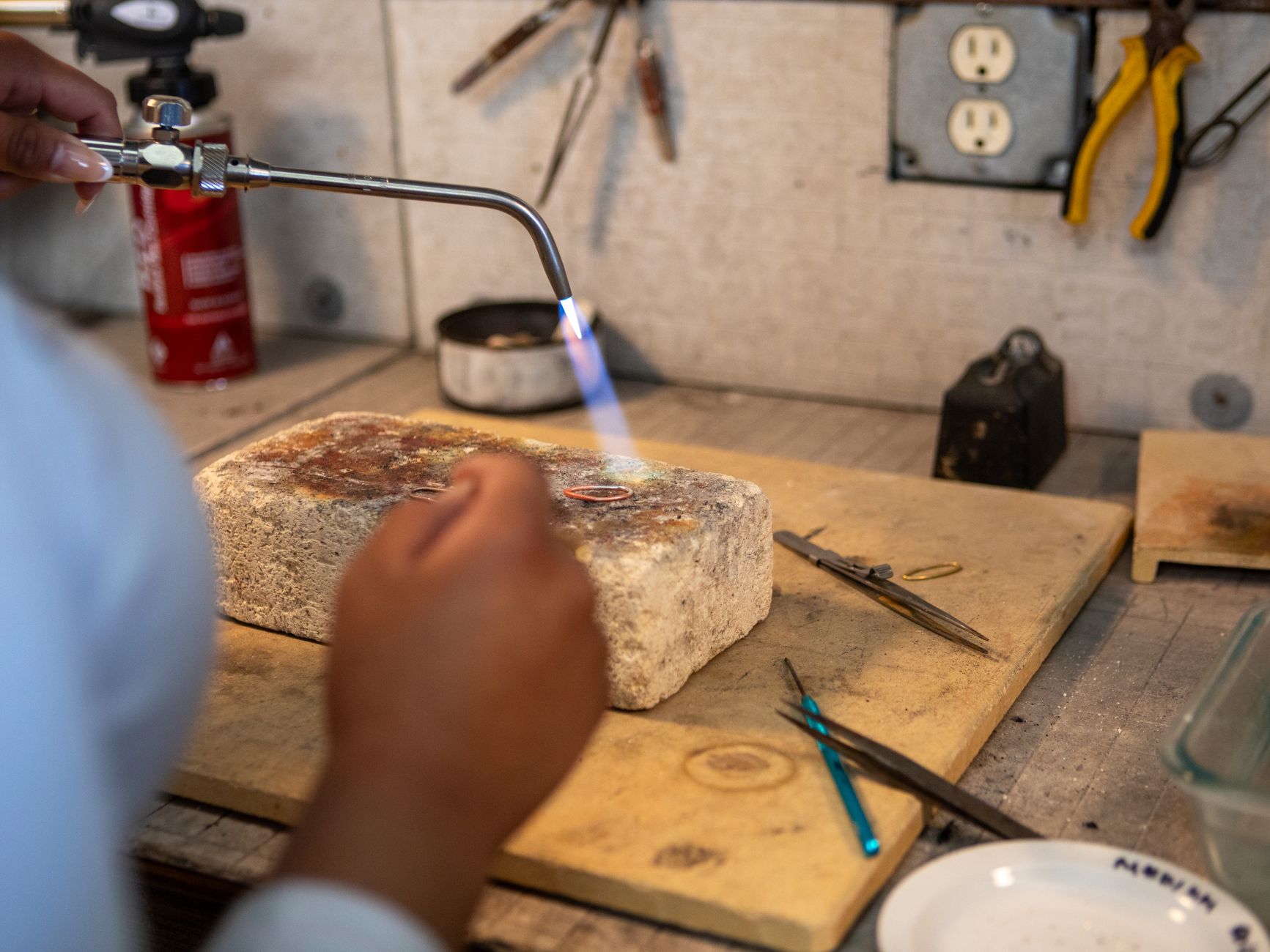 Close-up of hands using a blowtorch in a jewelry workshop. Tools and workspace visible.