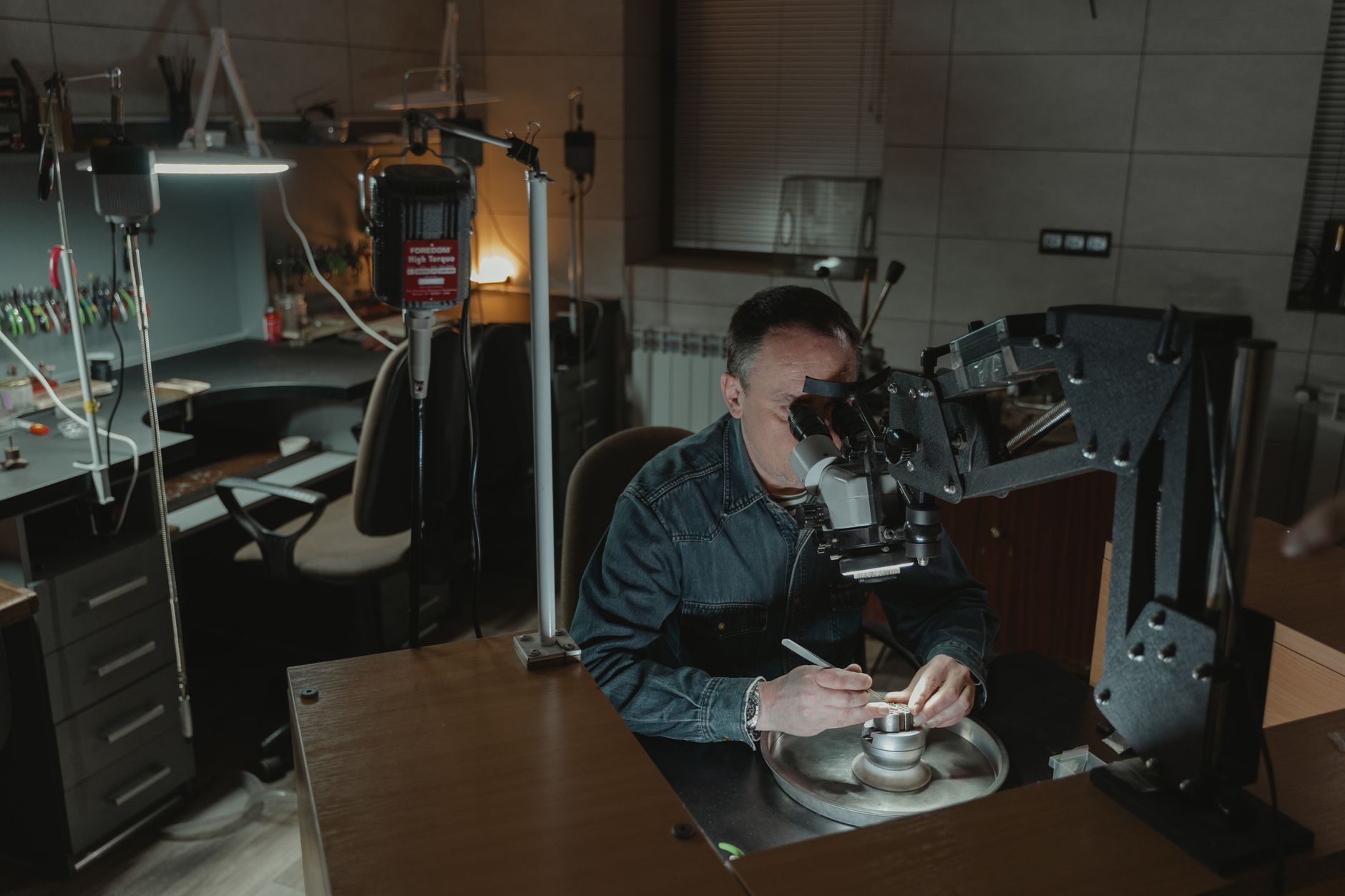Jeweler focusing on intricate work using a microscope in a dimly lit workspace.