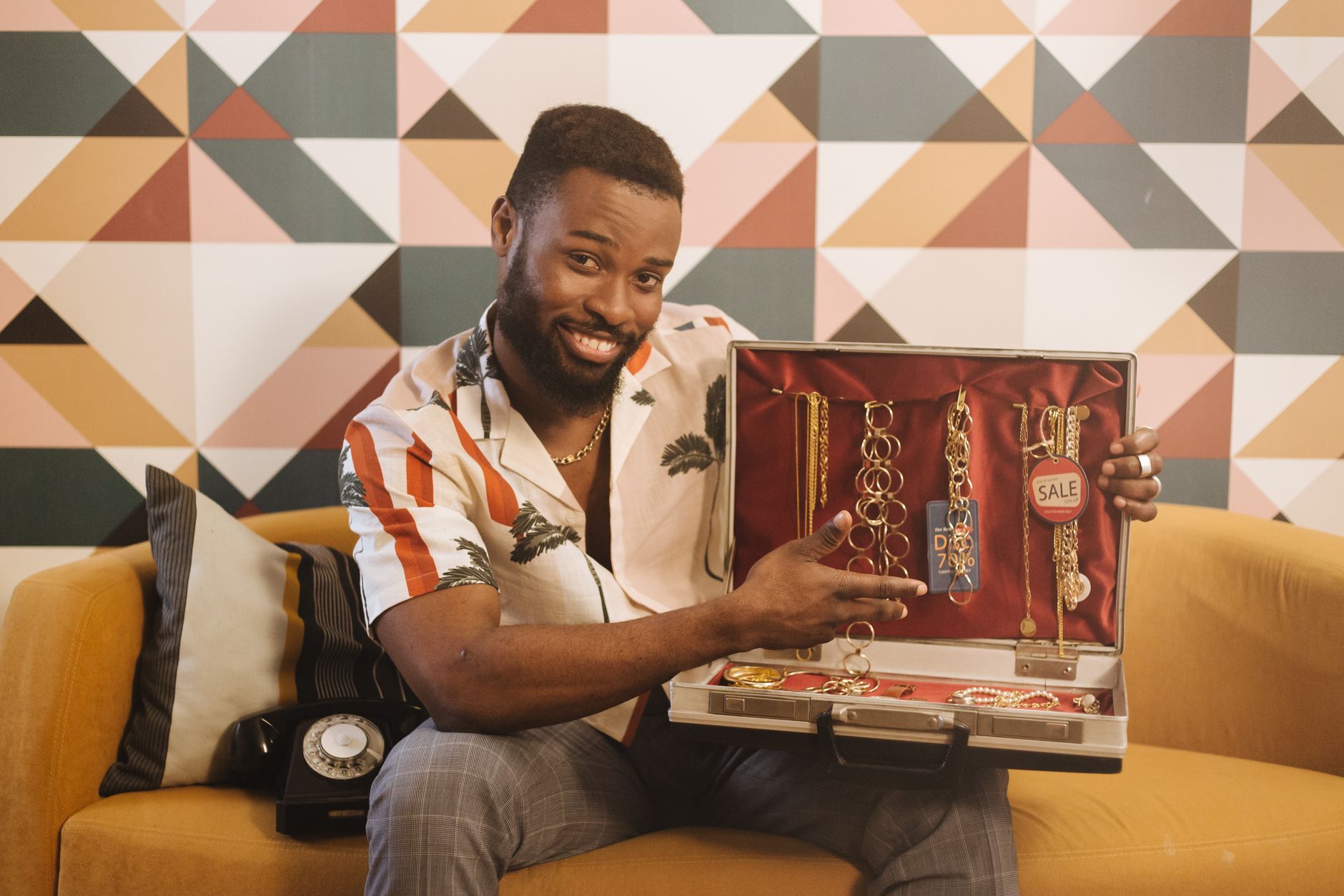 Smiling man sitting on a couch shows a variety of jewelry in a briefcase indoors.