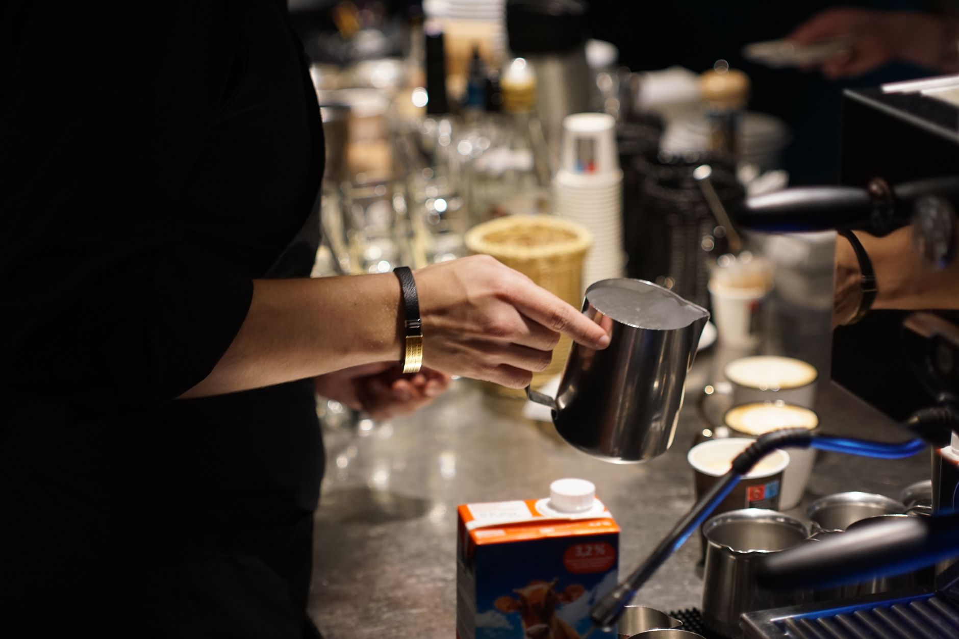 Close-up of a barista holding a pitcher in a bustling cafe setting, surrounded by cups and equipment.