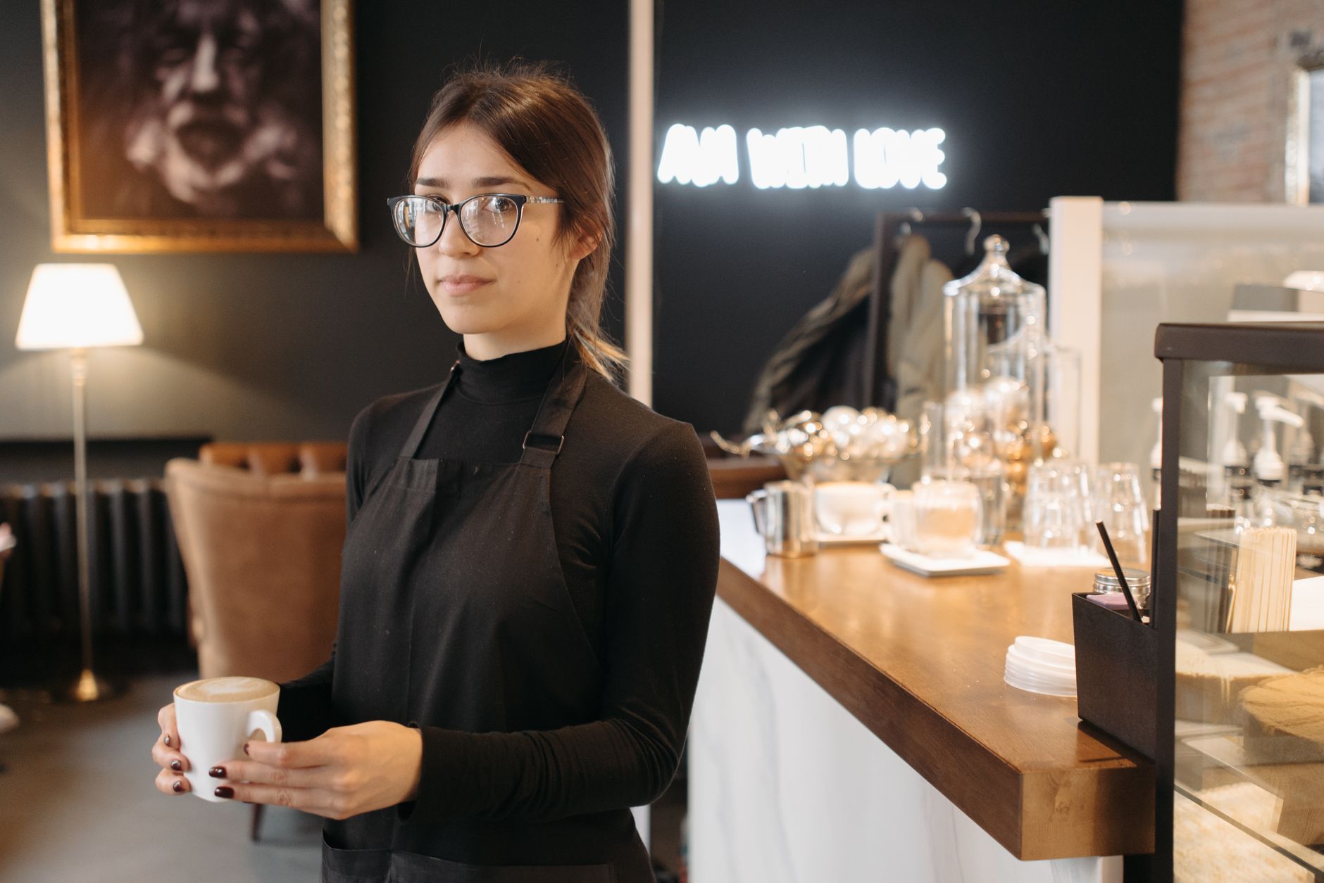 A barista holding a cup of coffee in a cozy café with warm lighting and stylish decor.