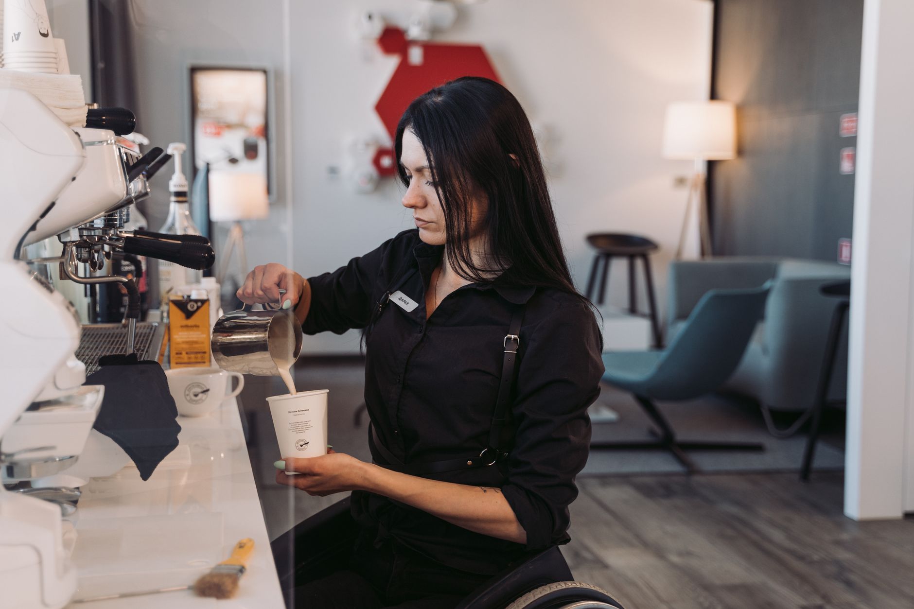 A woman in a wheelchair pours milk into a coffee cup at a modern cafe counter.