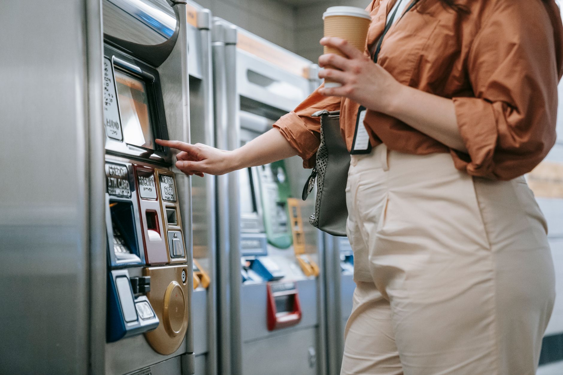 Woman purchasing subway ticket from a machine, holding a coffee cup.