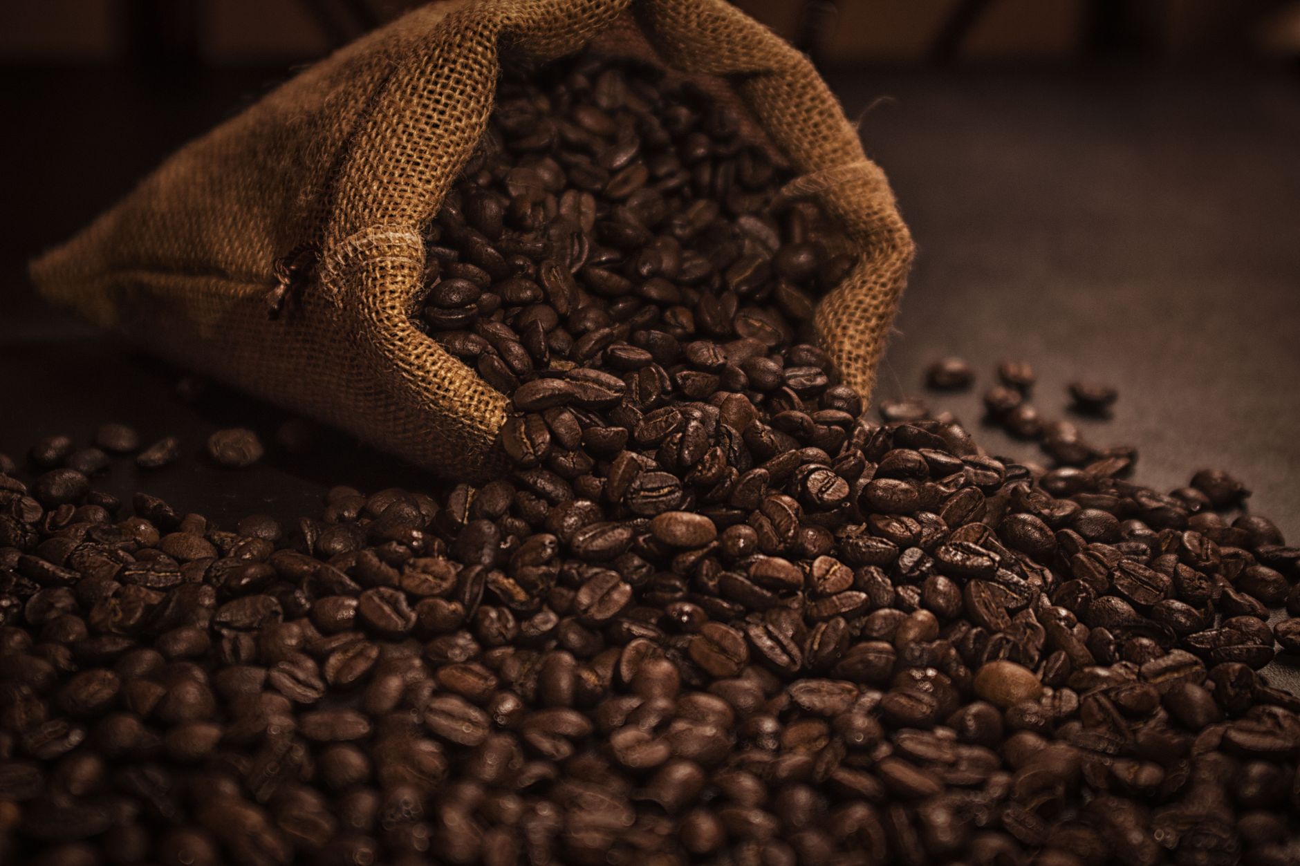 A burlap sack tipping over with dark roasted coffee beans spreading on a surface.