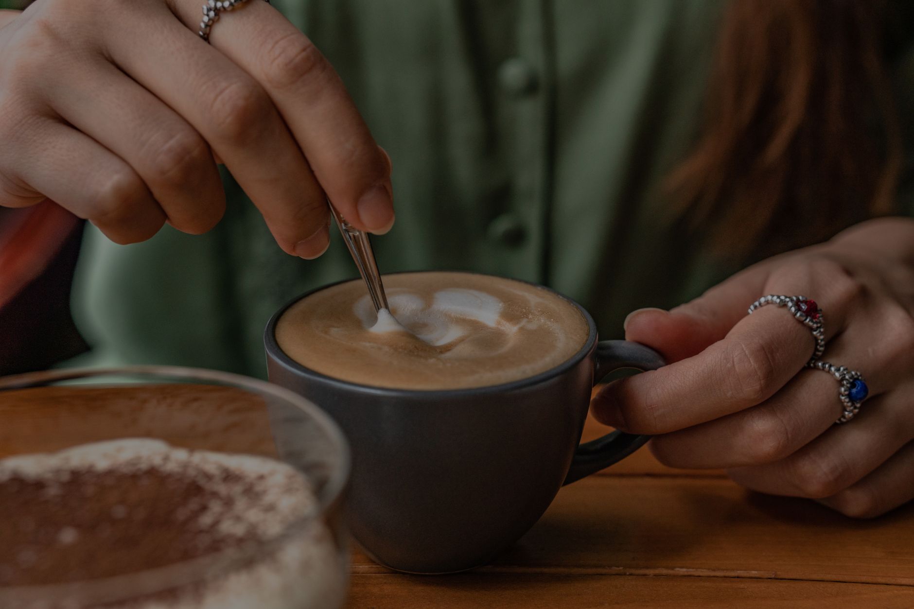 Hands stirring cappuccino in dark mug, cozy cafe setting.