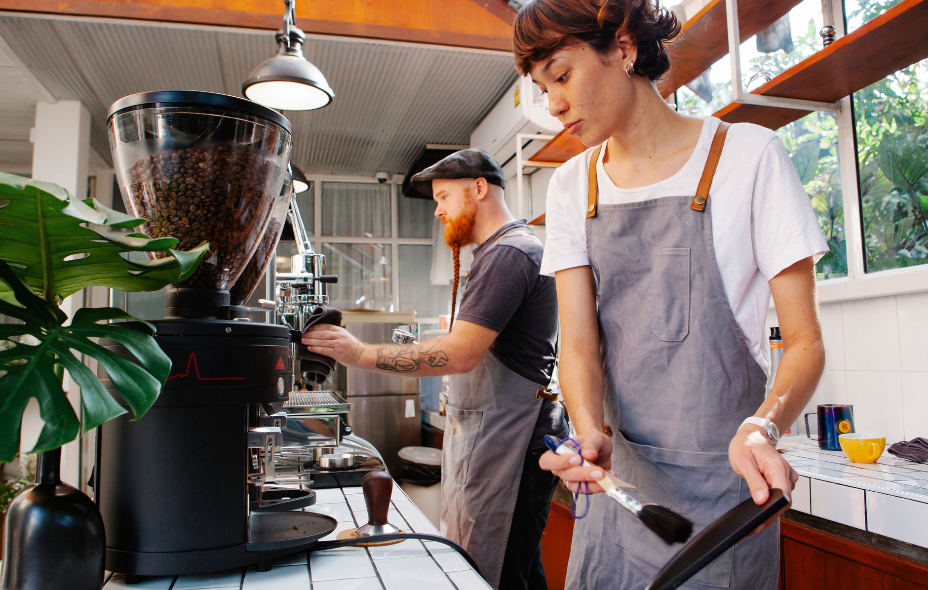 Crop female cafeteria employee with brush near bearded male partner against coffee machine working in daylight