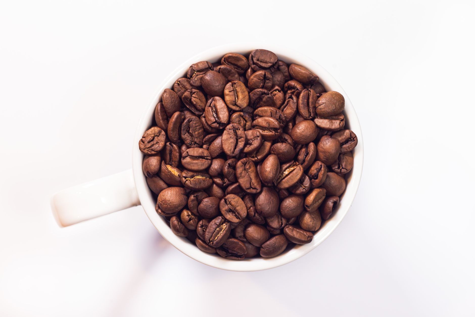 Freshly roasted coffee beans in a white mug, captured from above on a white background.