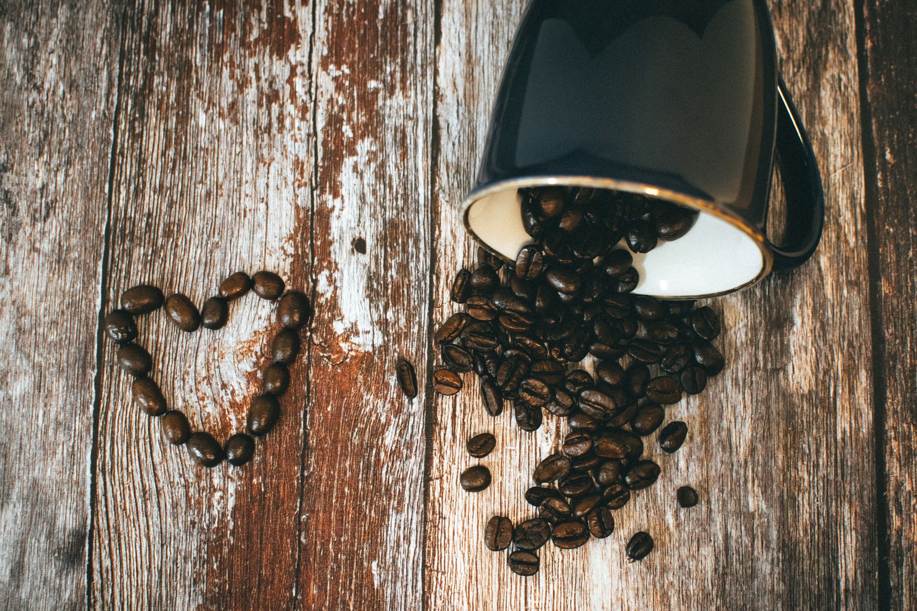 Dark roasted coffee beans spilled from mug on rustic wooden table with heart shape.