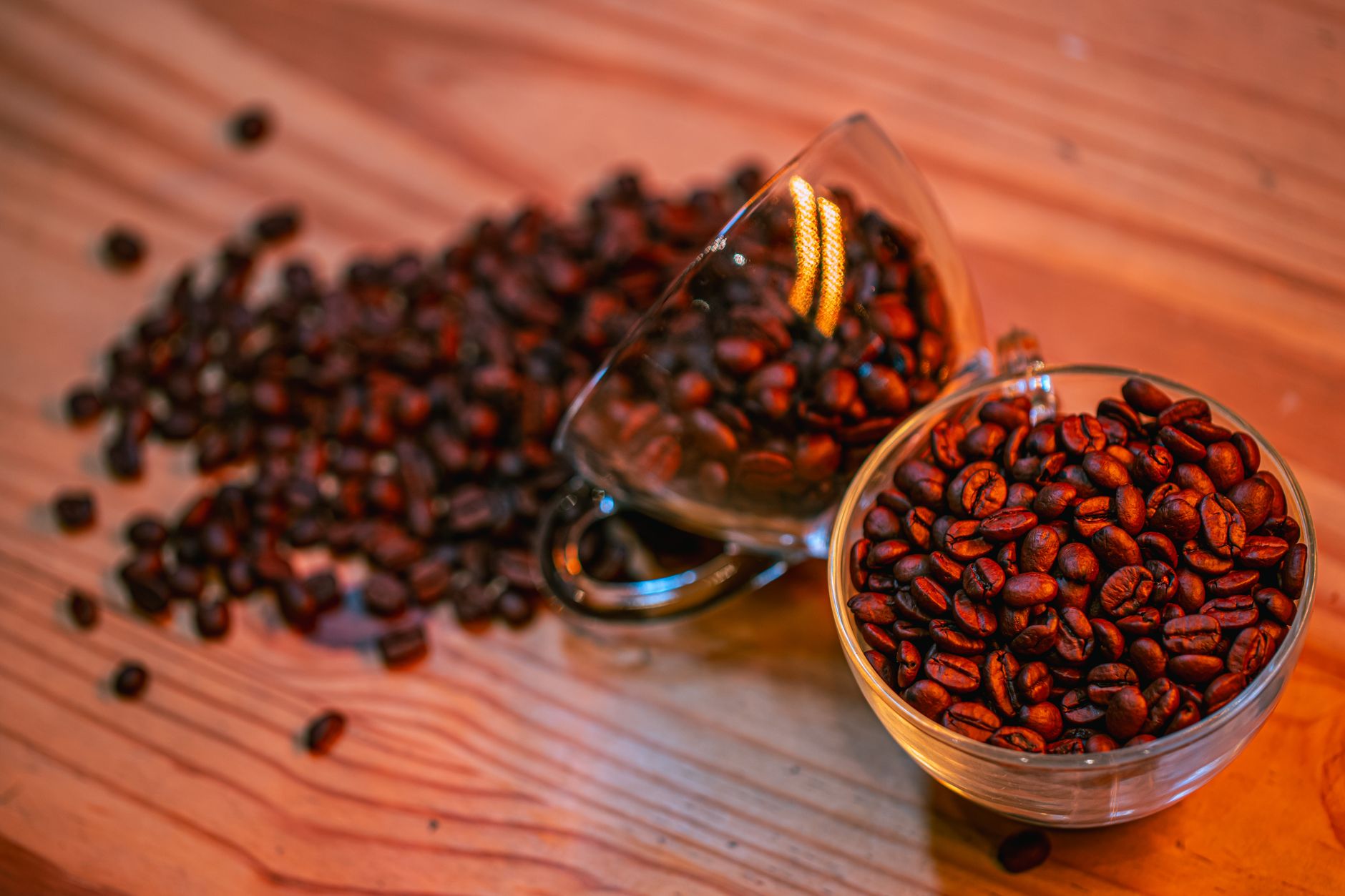 Close-up of rich brown coffee beans spilled from glass cups onto a wooden table.