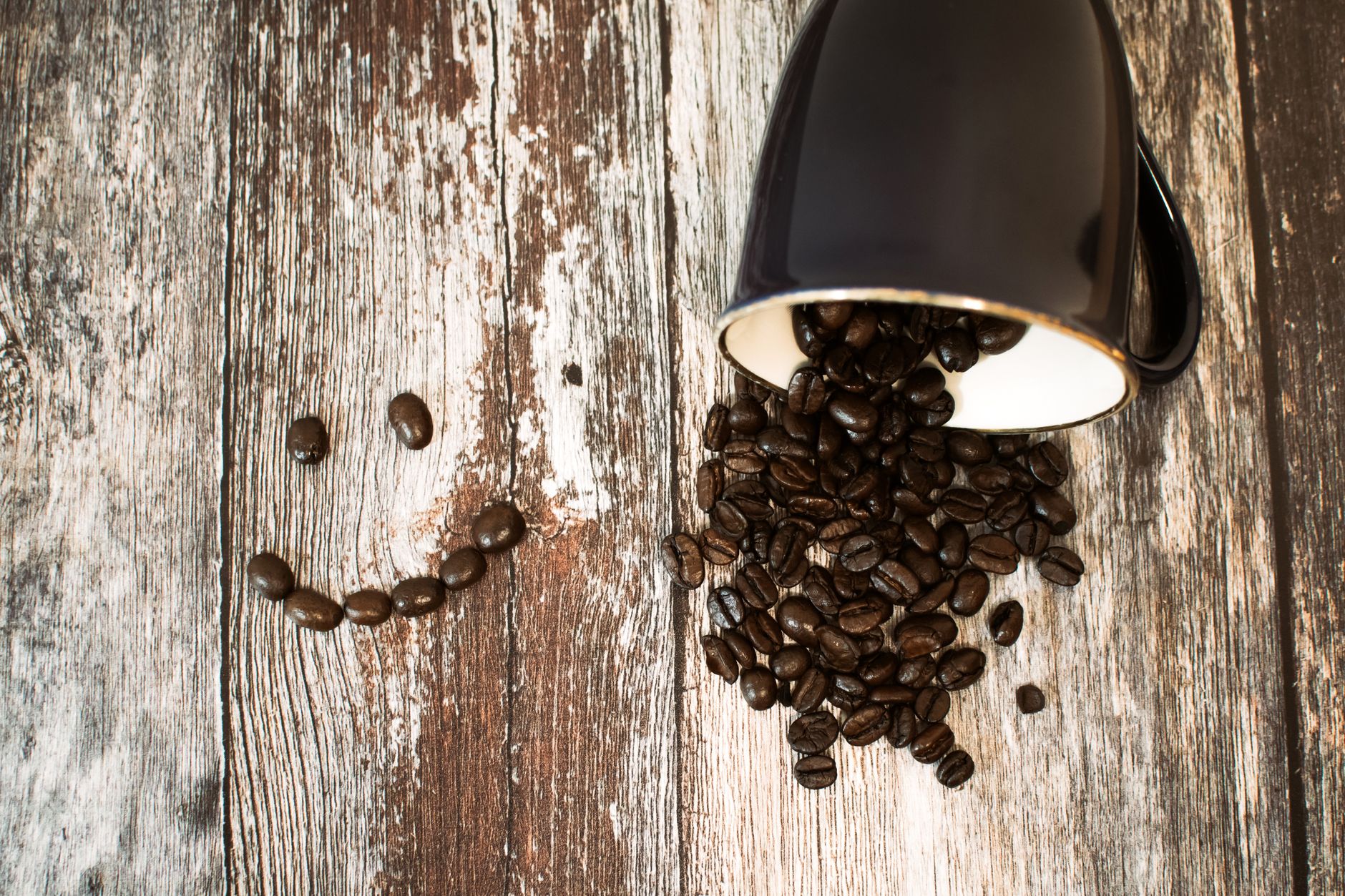 Smiley face of coffee beans beside a tipped mug on a wooden surface, evoking a rustic feel.