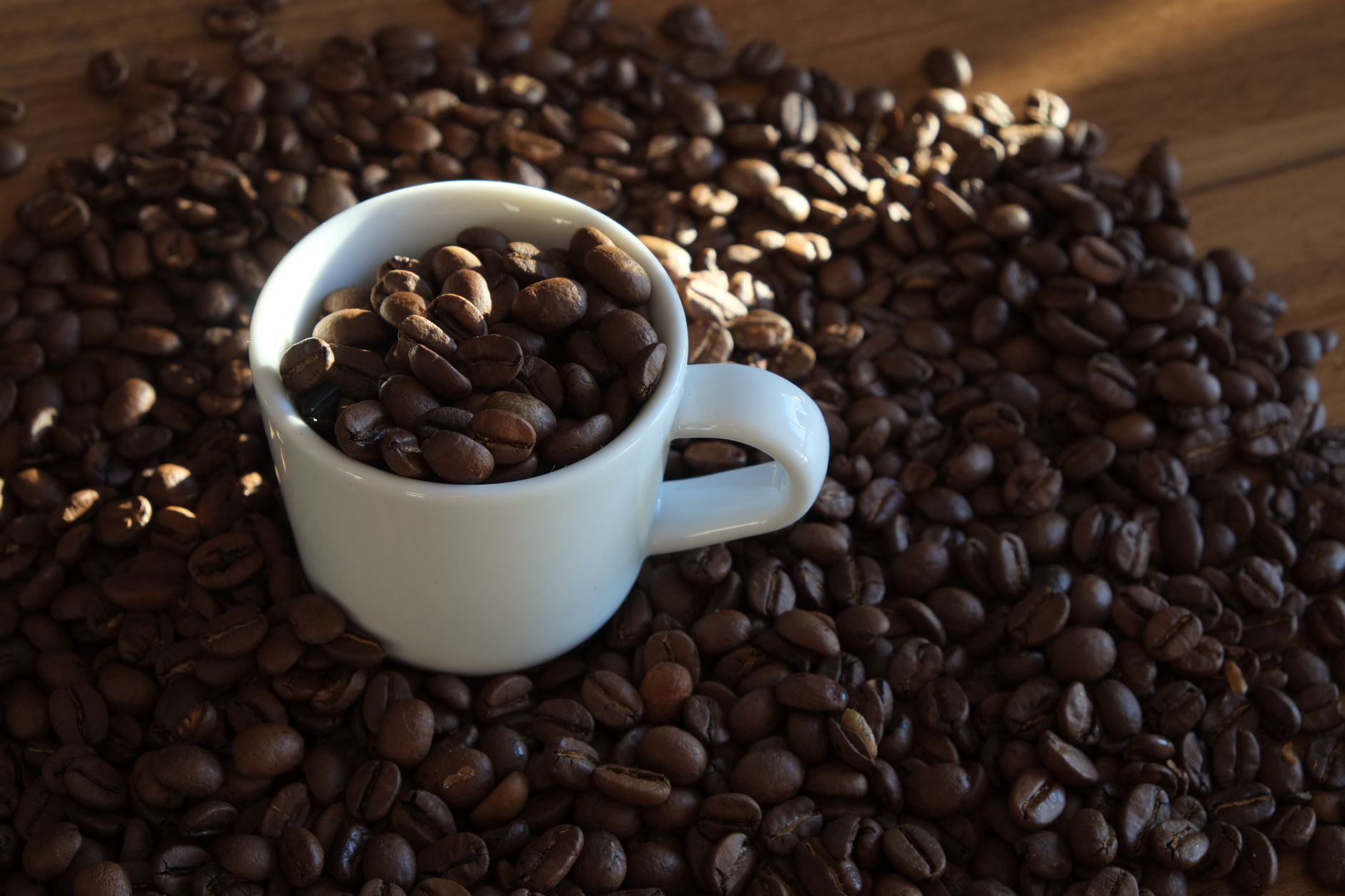 A white cup filled with coffee beans on a wooden table surrounded by scattered coffee beans.