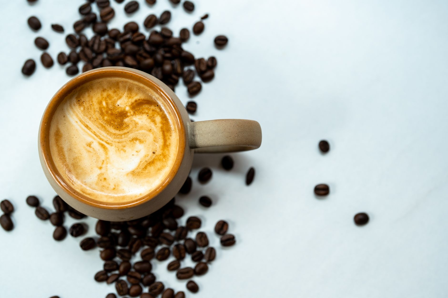 A top view of a coffee cup with frothy surface surrounded by roasted coffee beans.