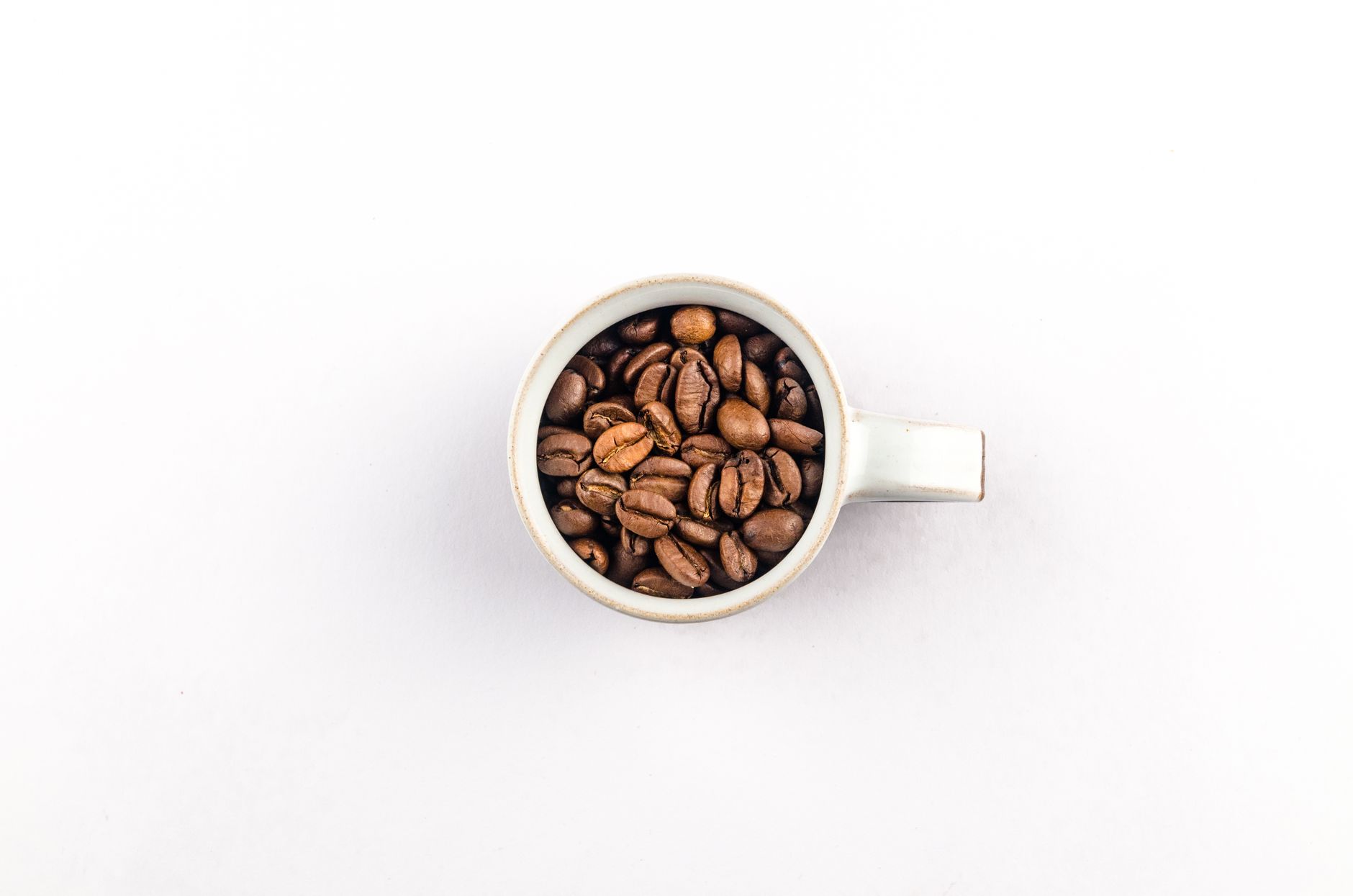 Overhead shot of roasted coffee beans filling a white ceramic cup against a plain background.
