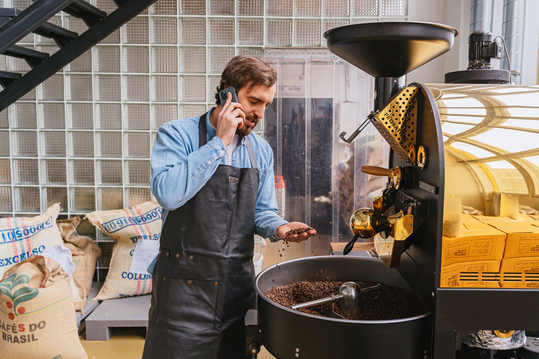 Man in apron roasting coffee beans while talking on the phone in a cafe setting.