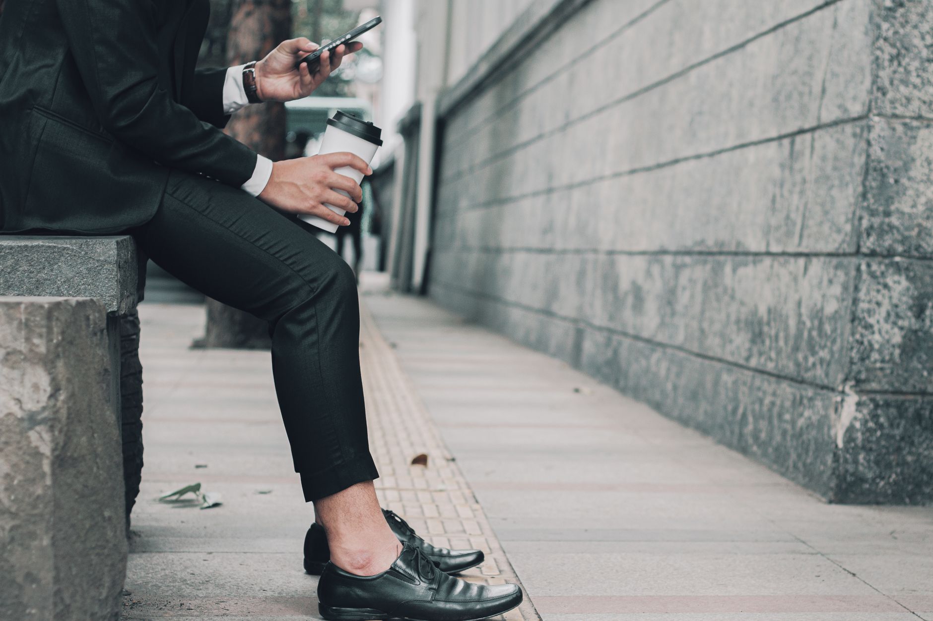 Stylish businessman in black suit sitting on street curb, holding coffee, using smartphone.