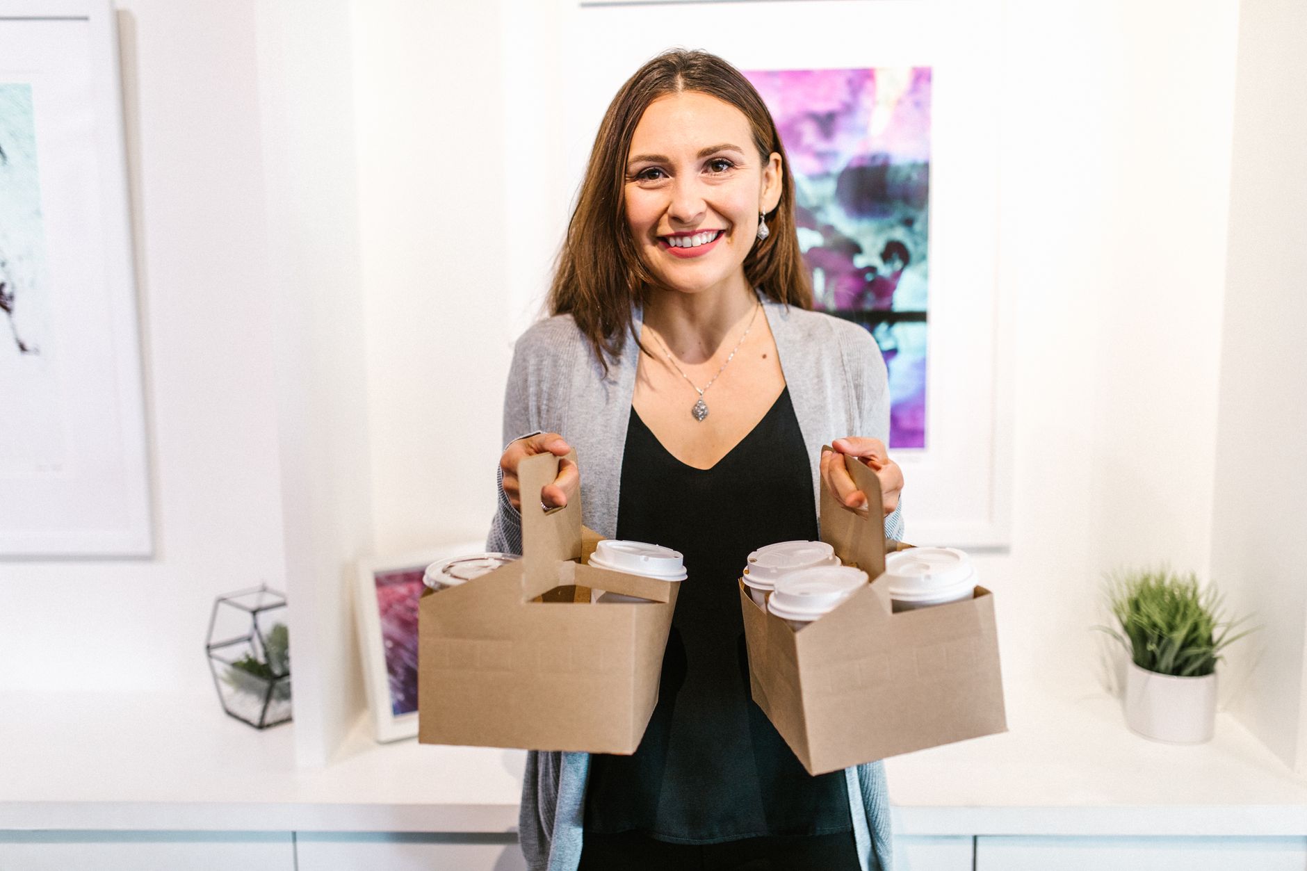 A cheerful woman holding two cardboard carriers with coffee cups, smiling indoors.