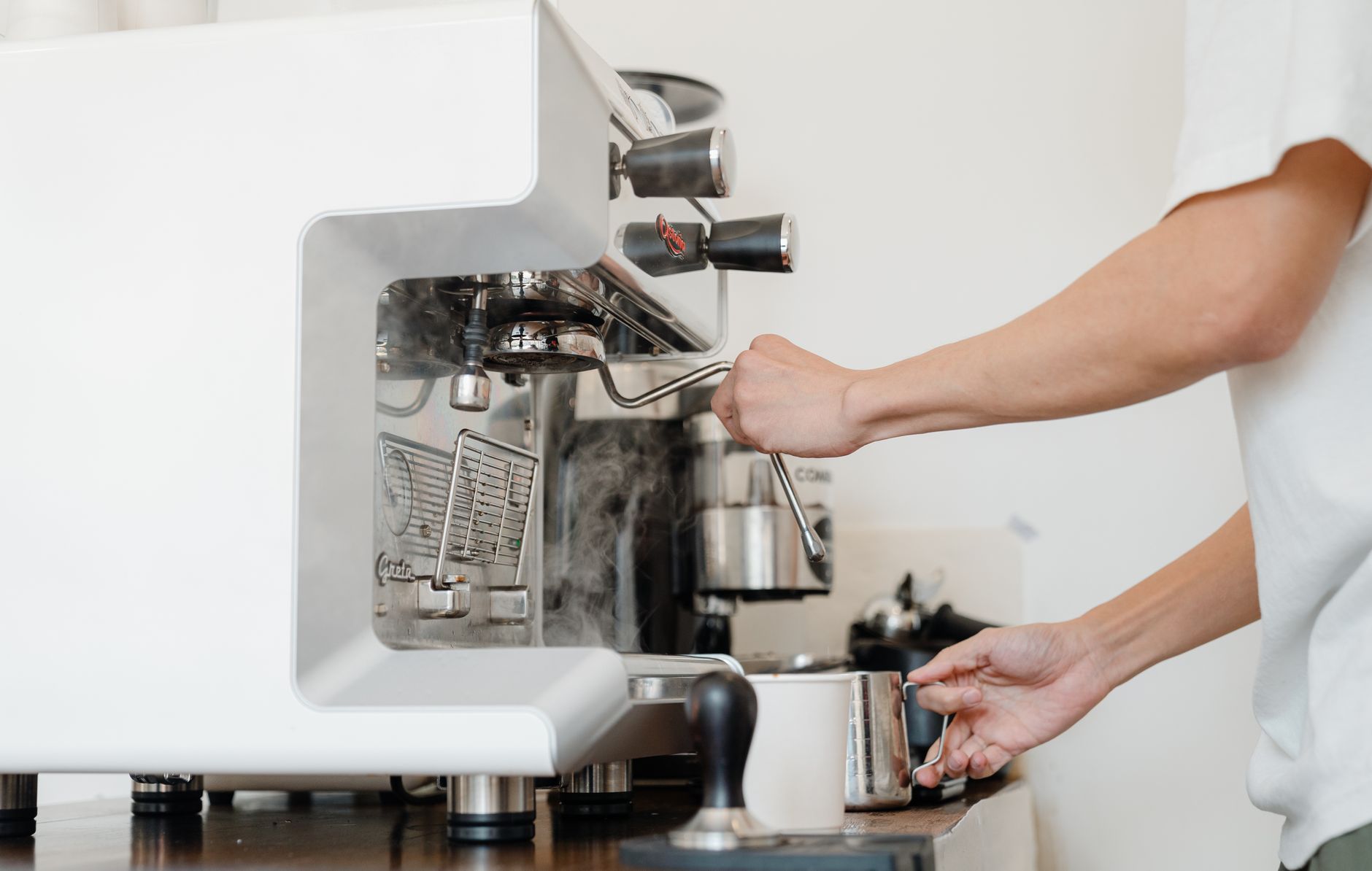 Side view crop anonymous barista in white shirt preparing to steam milk in frothing pitcher using professional coffee machine steam wand