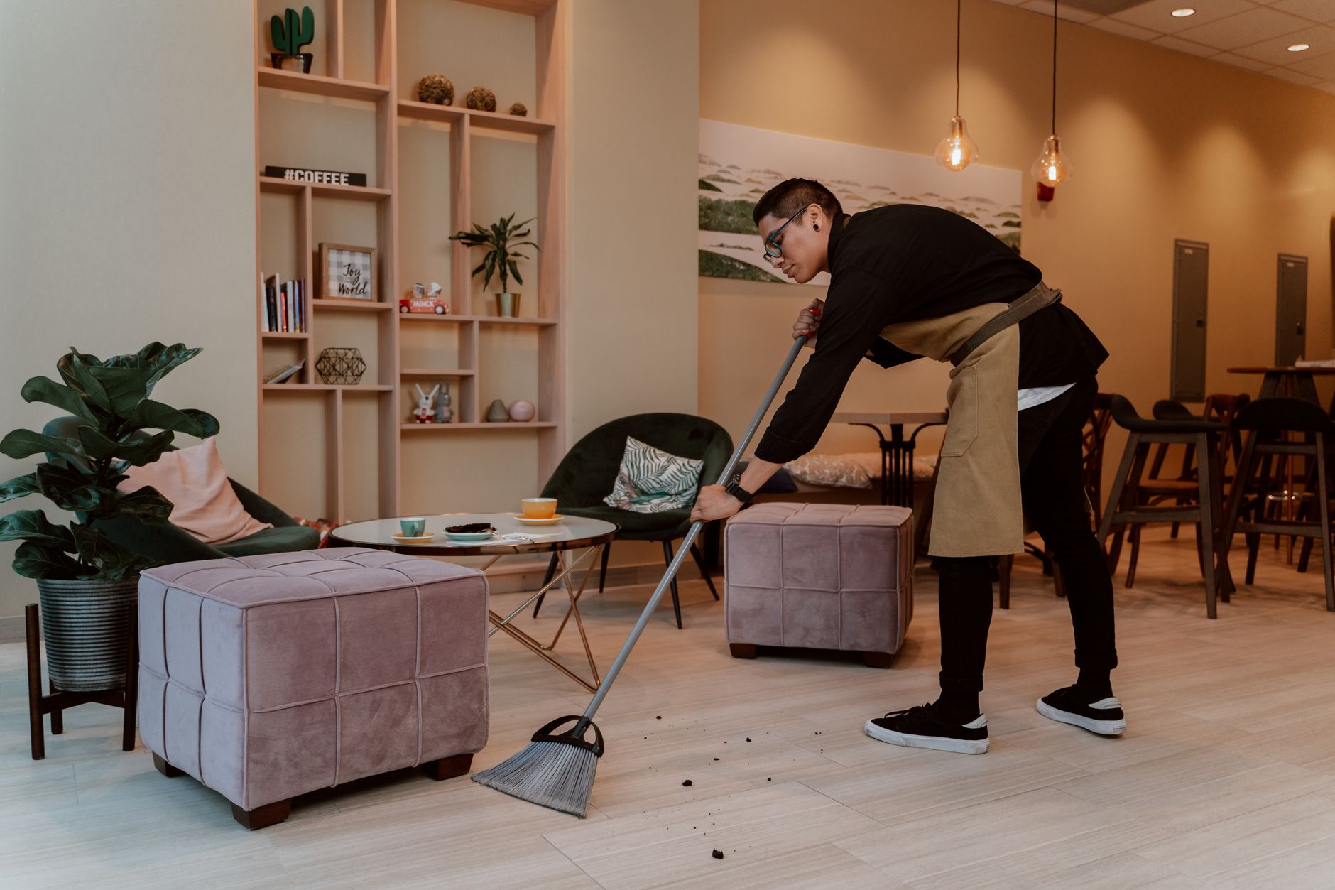 A barista sweeping the floor in a cozy, stylish coffee shop with modern decor and seating.